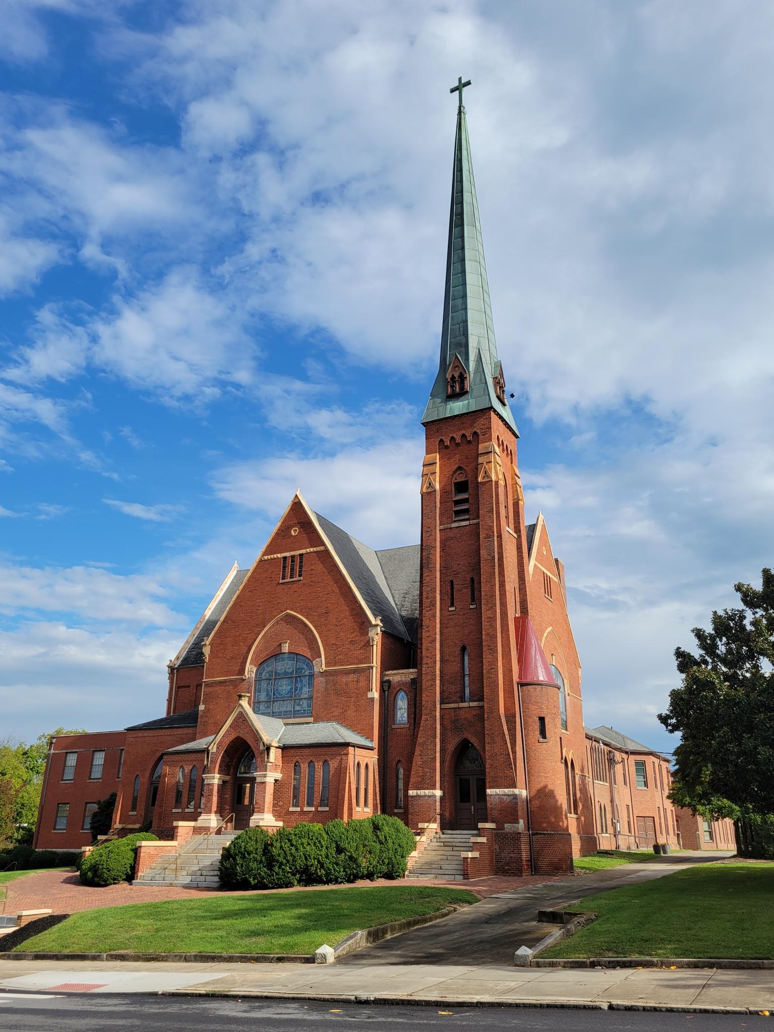 Large brick church with a tall green steeple under a bright, cloudy sky.