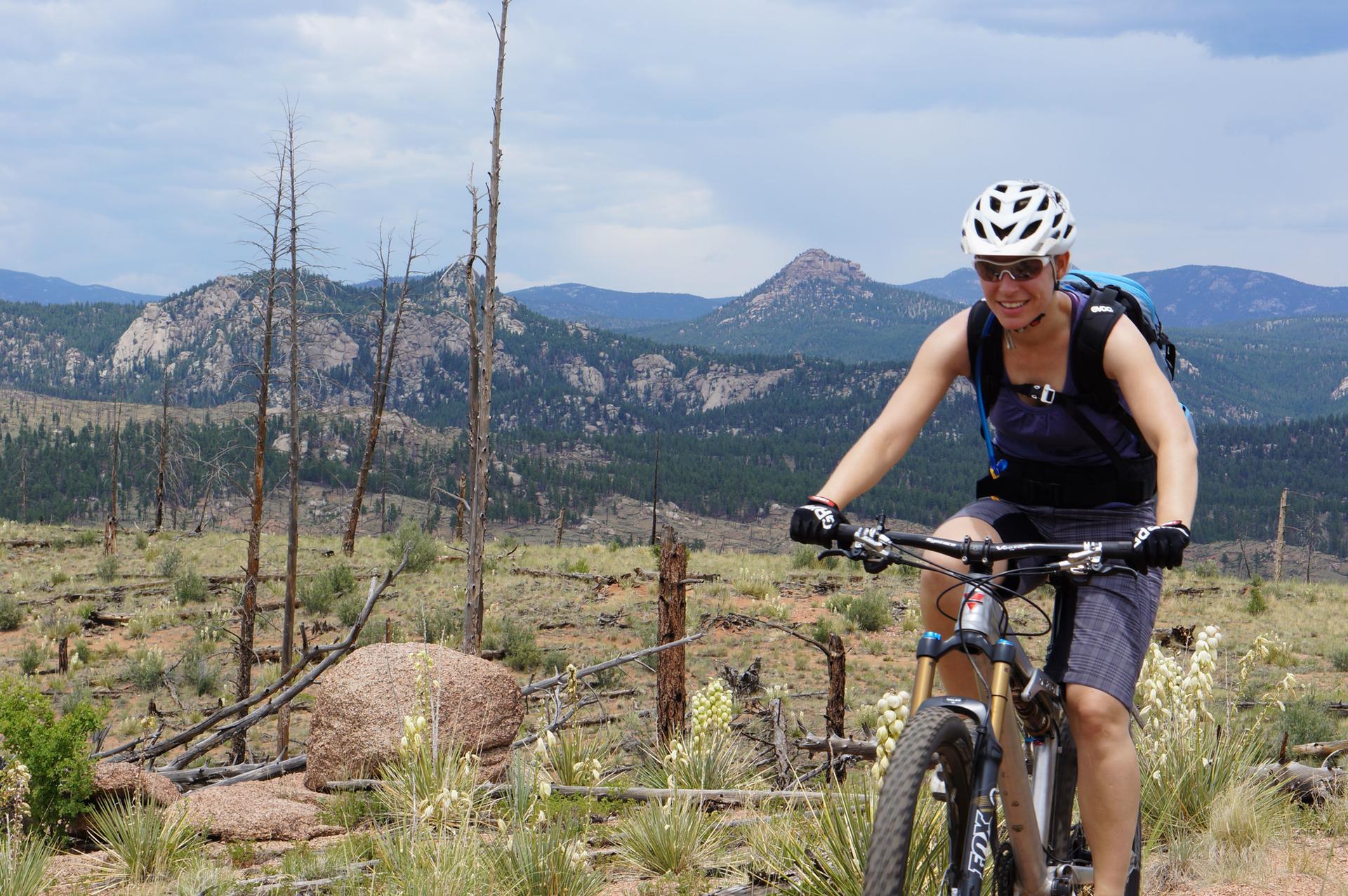 A smiling woman mountain bikes on a trail with mountains in the background.