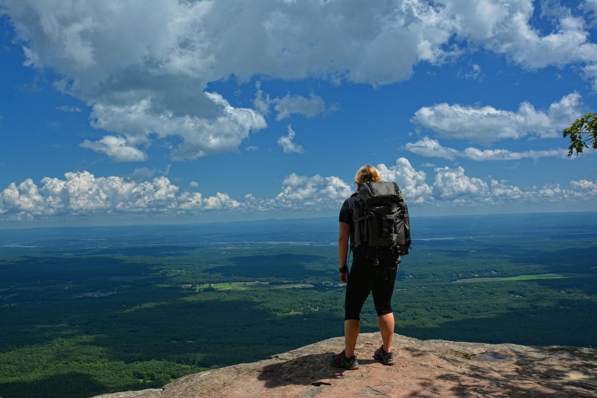 Hiker with backpack overlooking a vast forested landscape under cloudy sky.