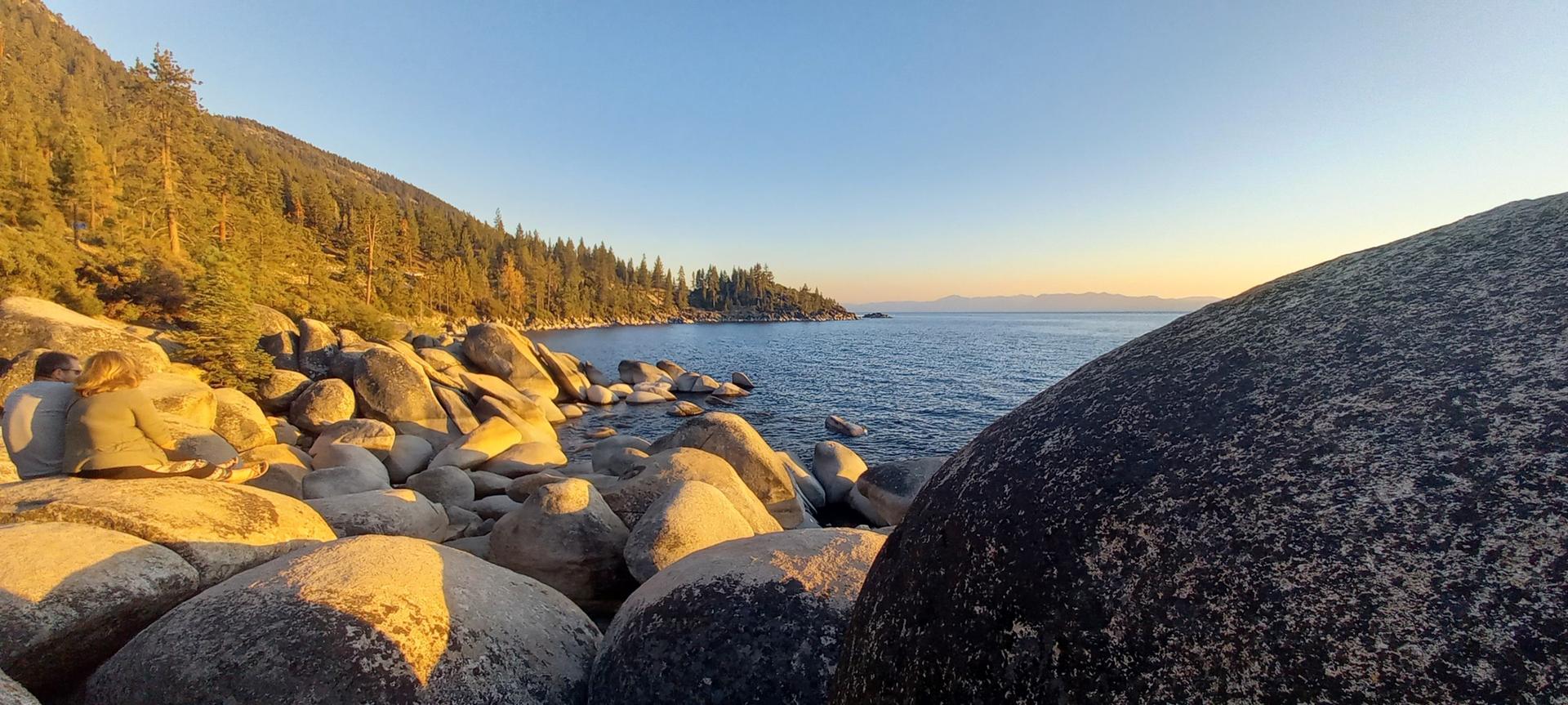 Golden hour light illuminates a rocky shoreline and calm lake with forested hills.