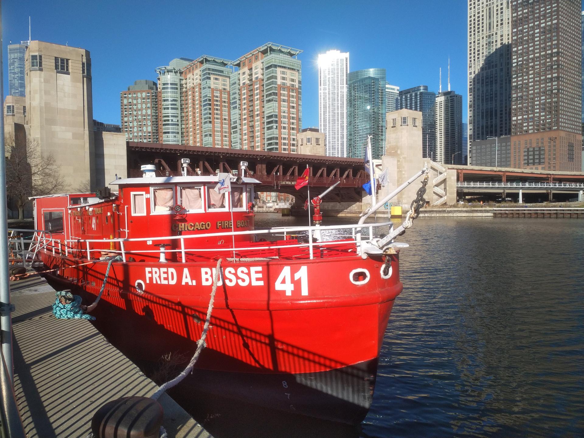 A bright red fireboat is docked by a river with tall city buildings.