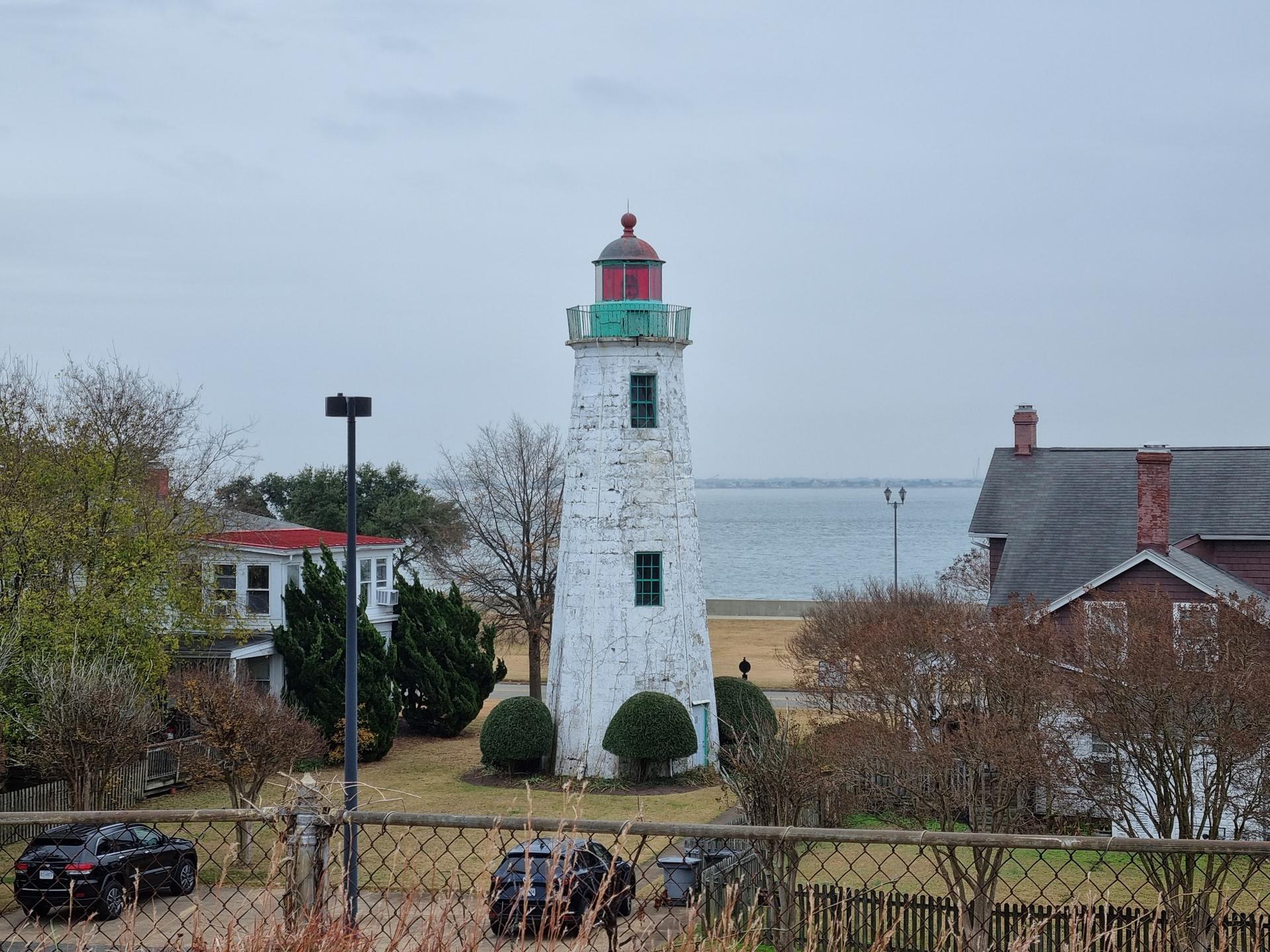 White lighthouse with red and green top, houses, and water under overcast sky.