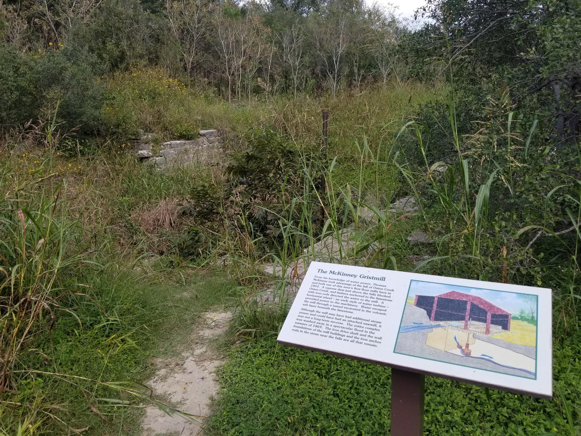 Informational sign on a dirt path next to overgrown ruins in a field.