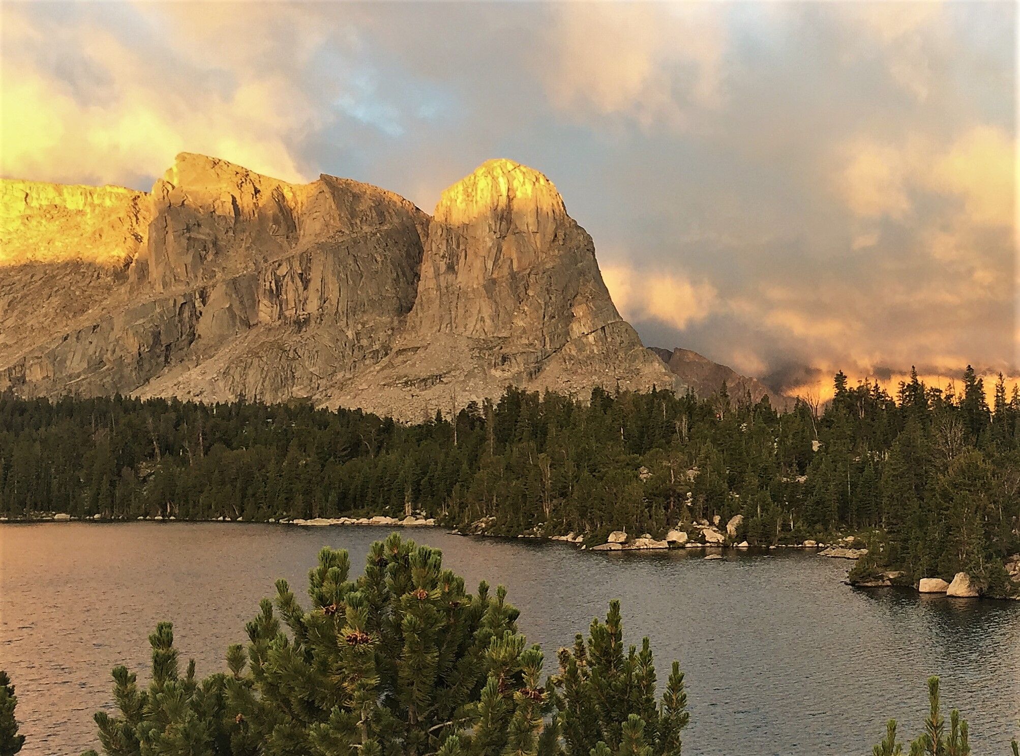Golden light illuminates rugged mountains above a forested lake at sunset. - Bears Ears Trail