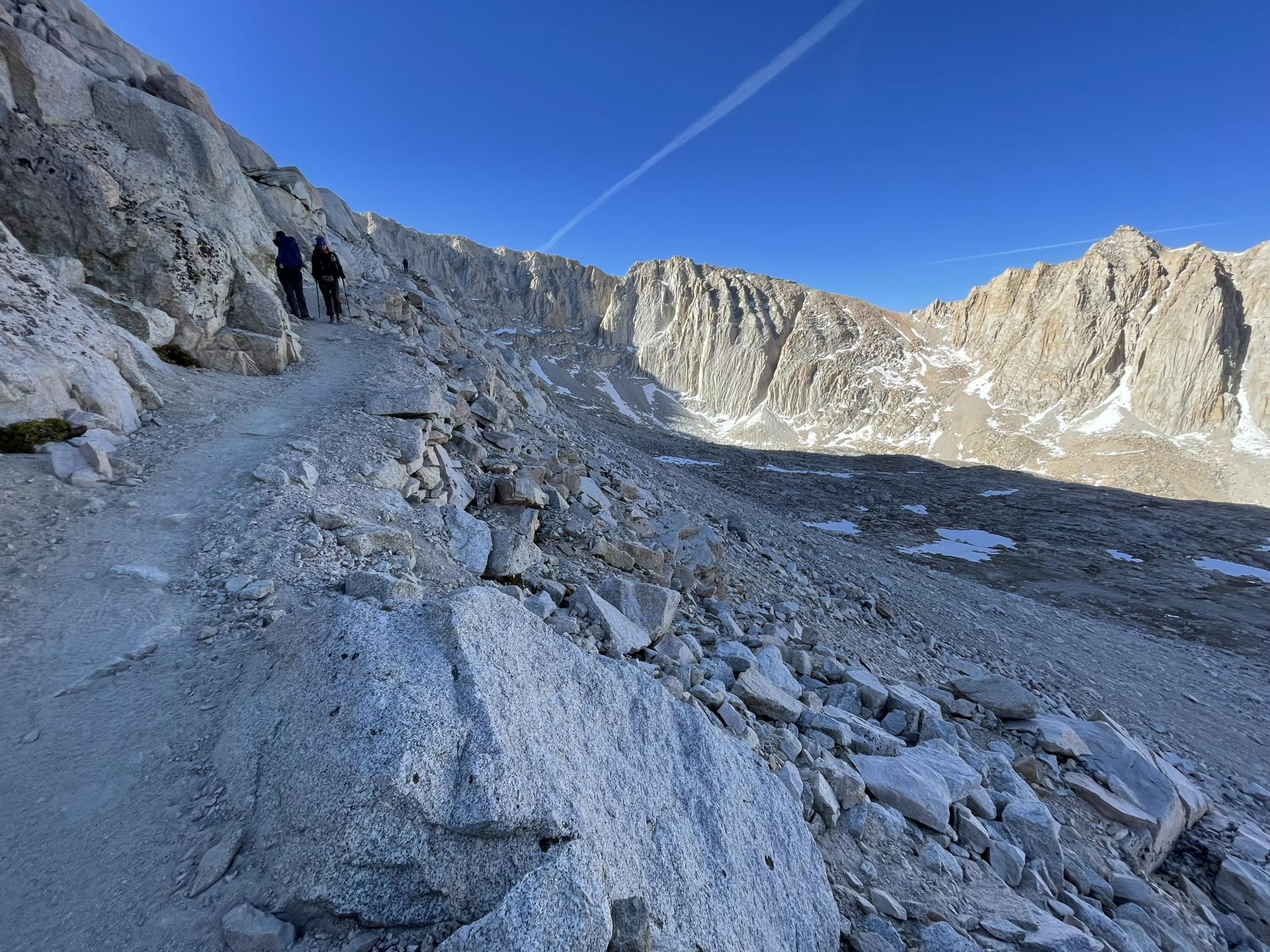 Two hikers on a rocky mountain trail overlooking a vast, snowy valley.