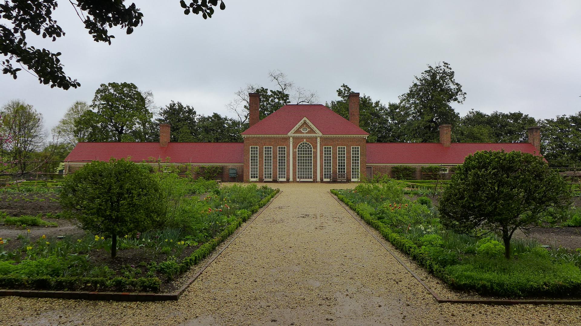 Large brick building with red roof, surrounded by formal gardens on a cloudy day.