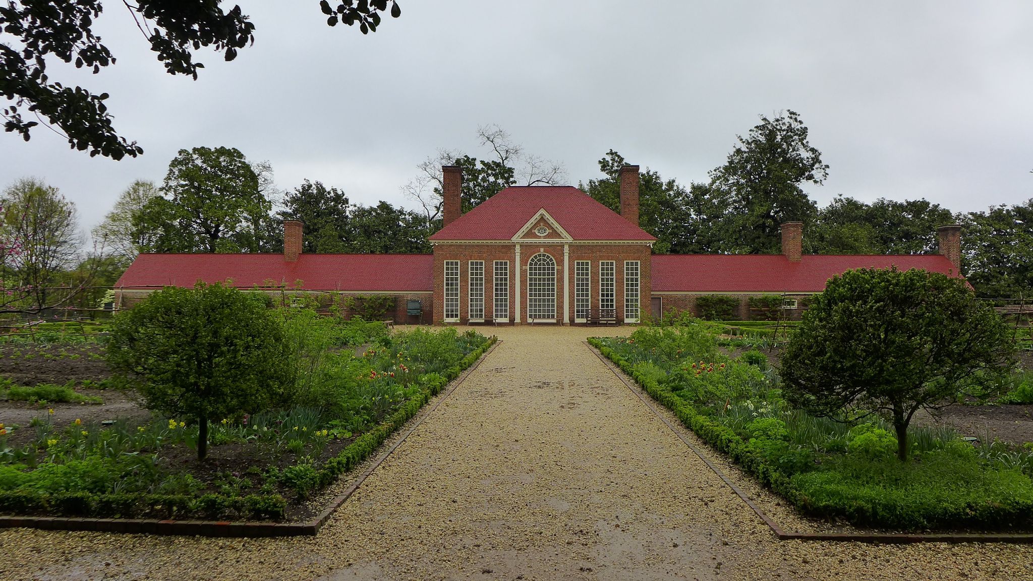 Large brick building with red roof, surrounded by formal gardens on a cloudy day. - Mount Vernon Trail