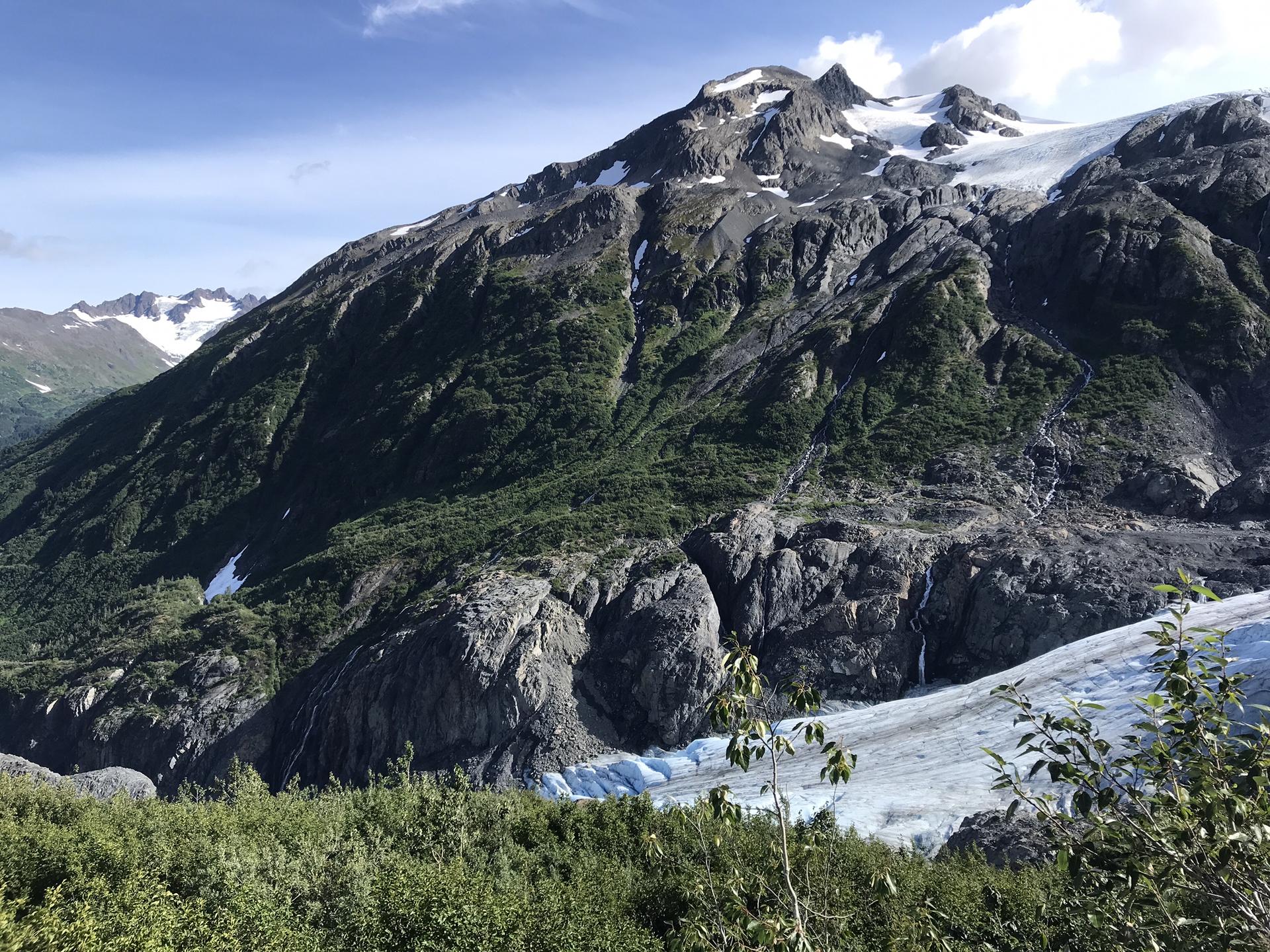 A large green mountain with a glacier at its base under a bright sky.