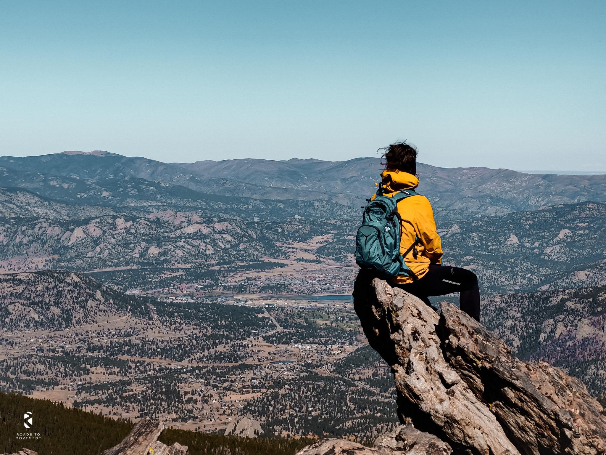 Person in yellow jacket and backpack sitting on a rock, overlooking mountains.