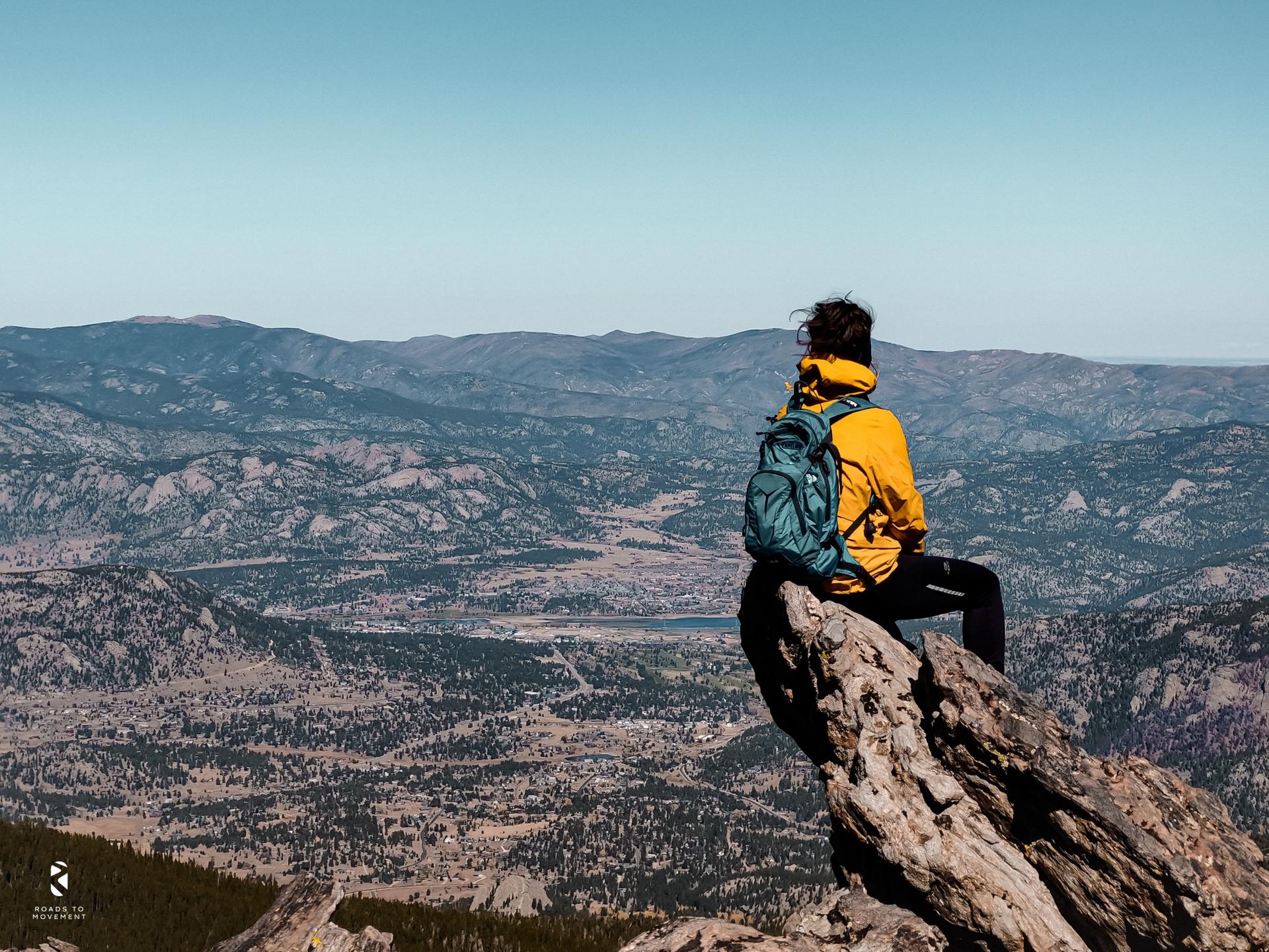 Person in yellow jacket and backpack sitting on a rock, overlooking mountains.