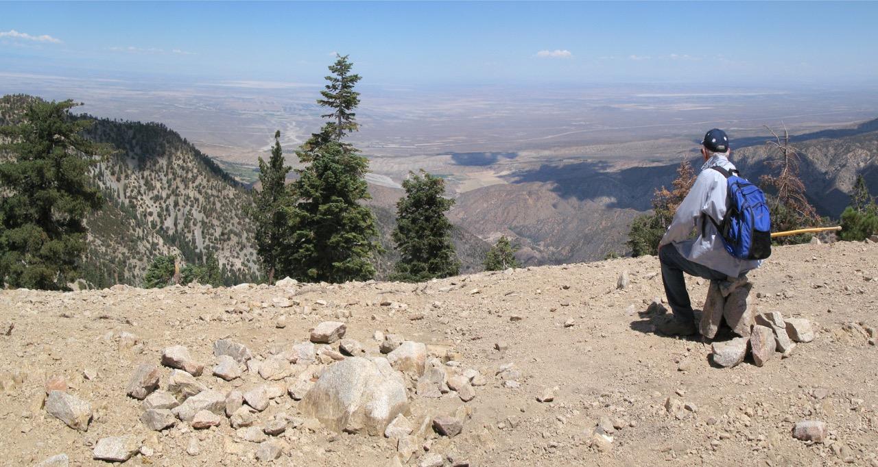 A person with a backpack sits, admiring a vast, arid mountain landscape.