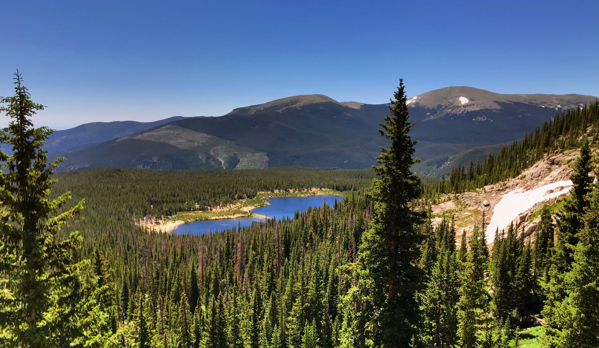 Vast mountain landscape with a clear blue lake surrounded by green pine trees. - Sandbeach Lake Trail