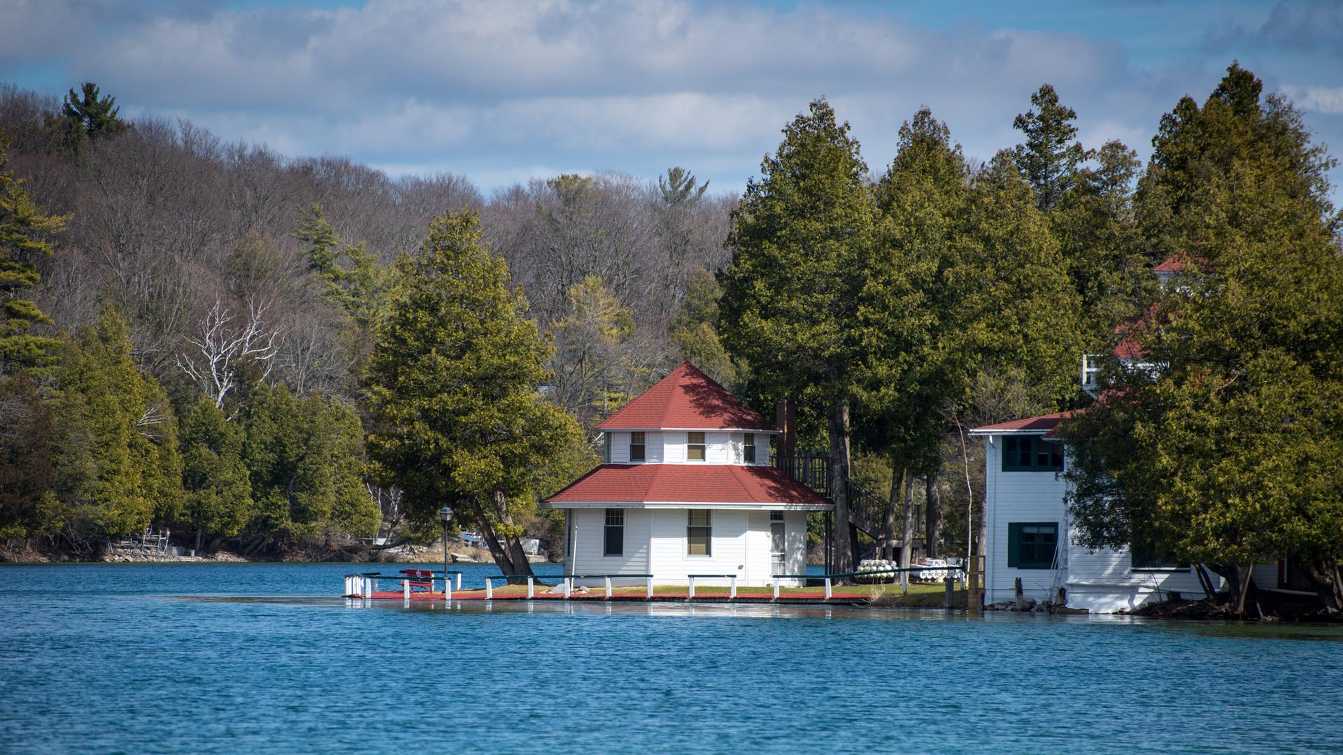 A white house with a red roof sits on a blue lake surrounded by trees.