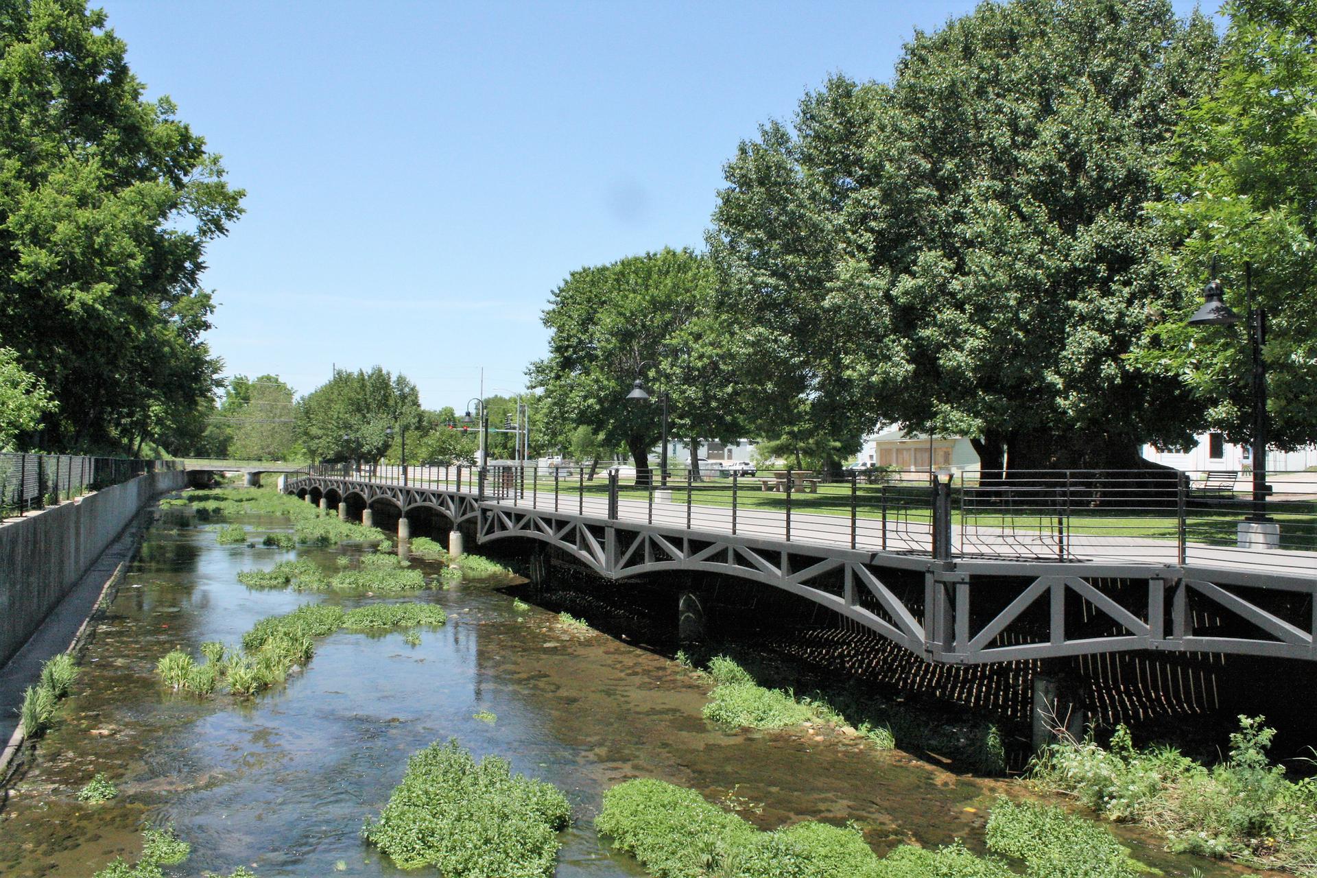 Sunny view of a pedestrian bridge over a narrow, vegetation-filled river with trees.
