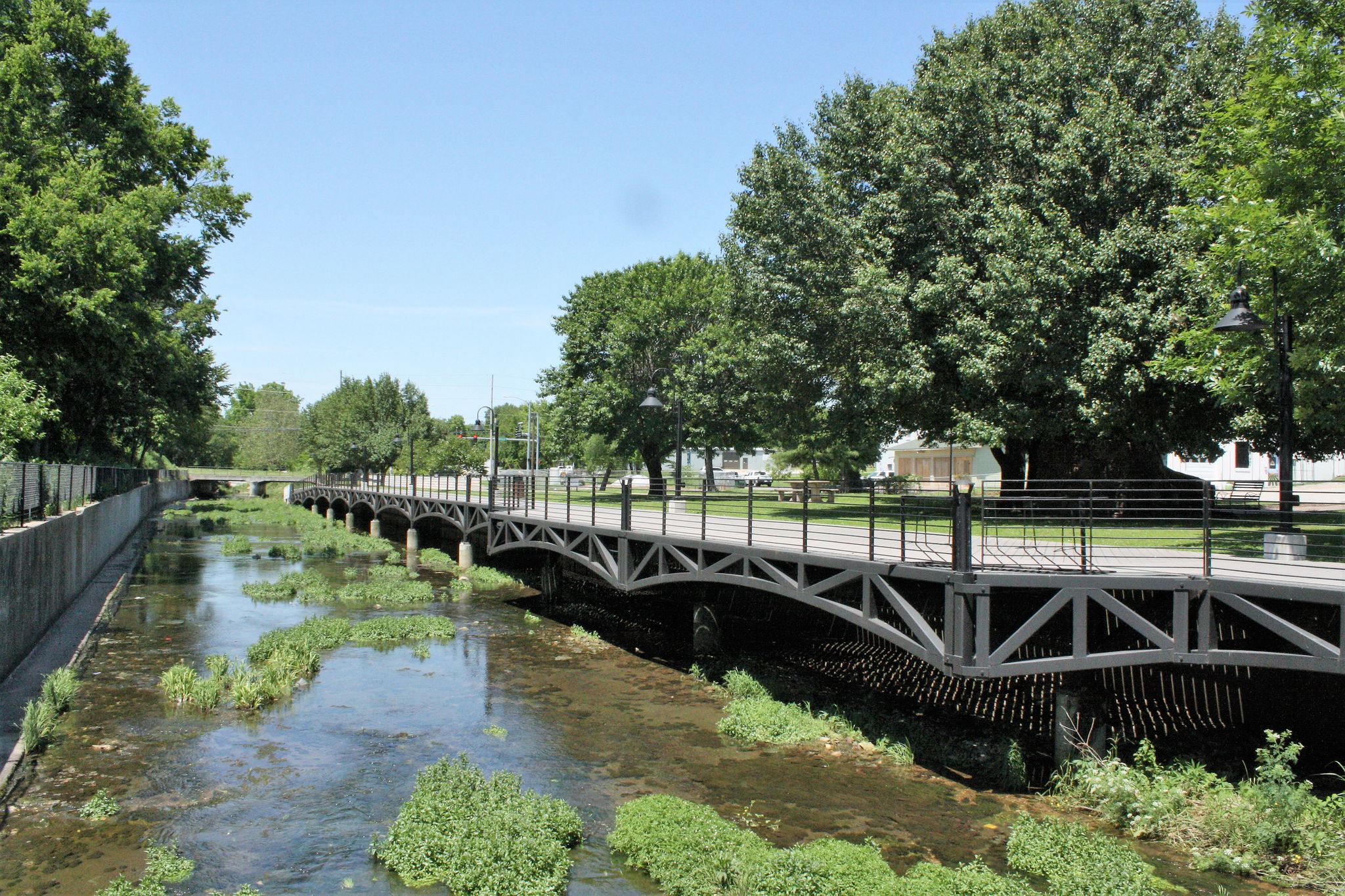 Sunny view of a pedestrian bridge over a narrow, vegetation-filled river with trees. - Razorback Regional Greenway – Boardwalk along Spring Creek loop from Pinnacle Open Space
