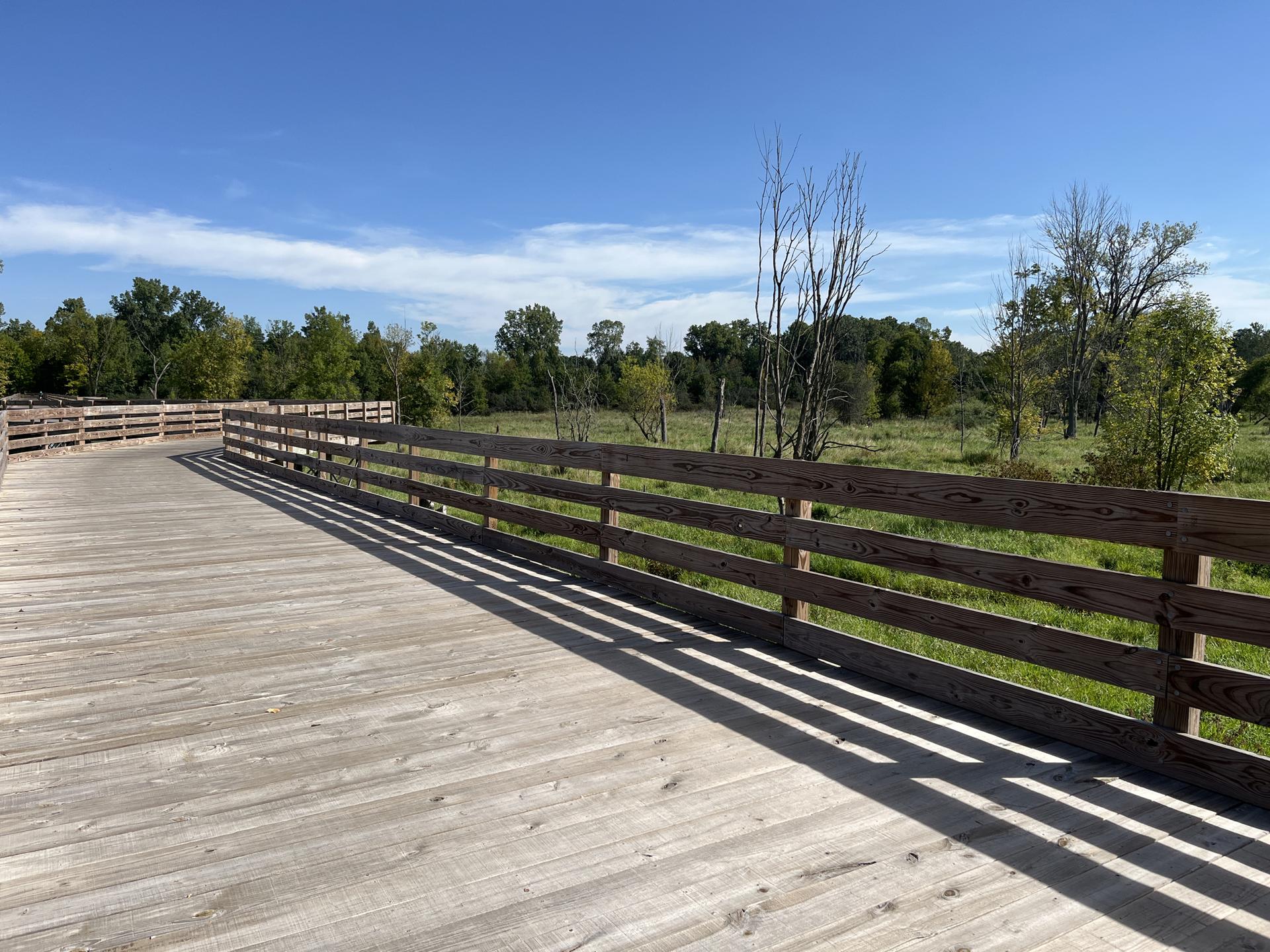 A long wooden boardwalk with railings overlooks a sunny, grassy field.