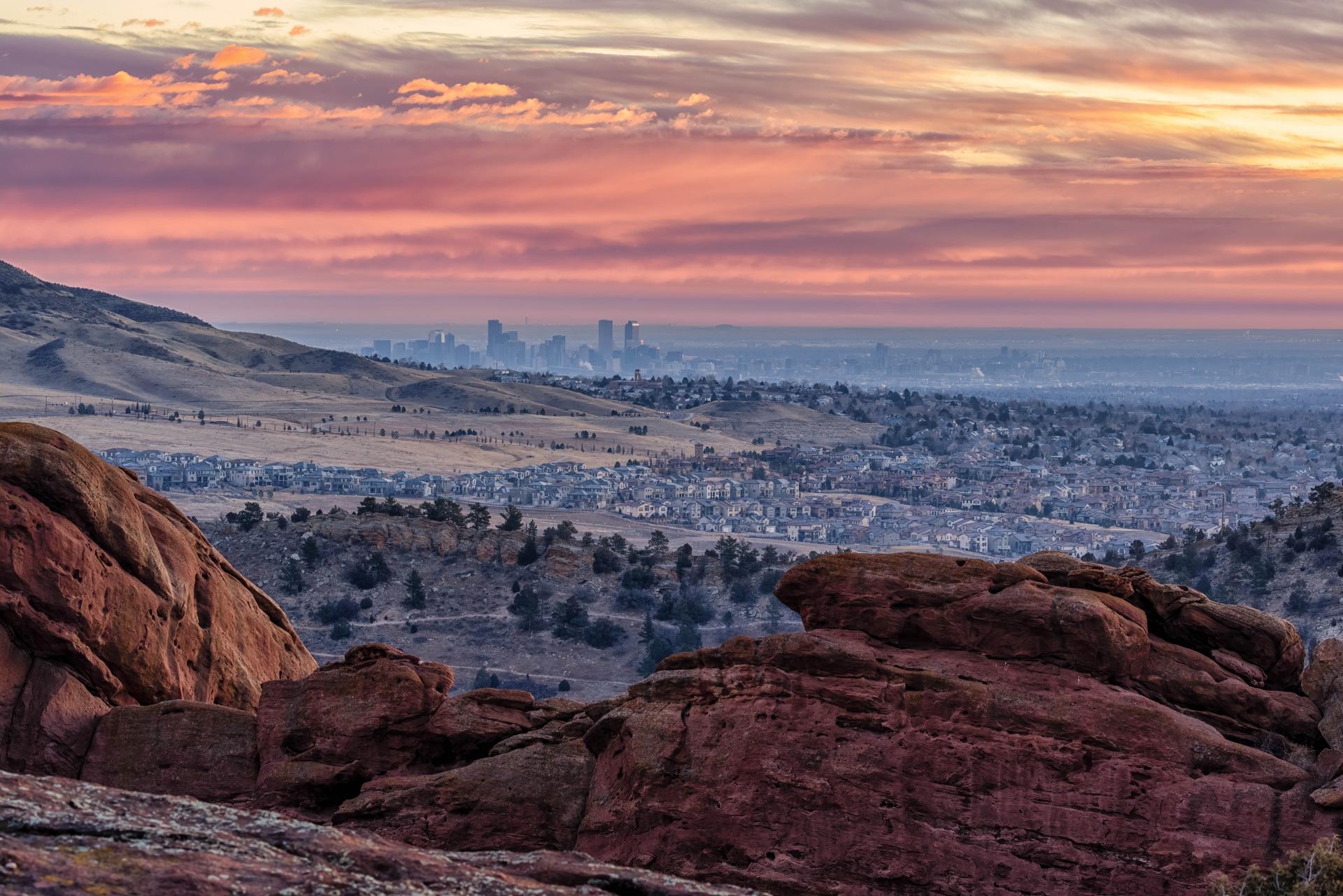 Distant city skyline at sunset, with colorful clouds and red rock foreground.