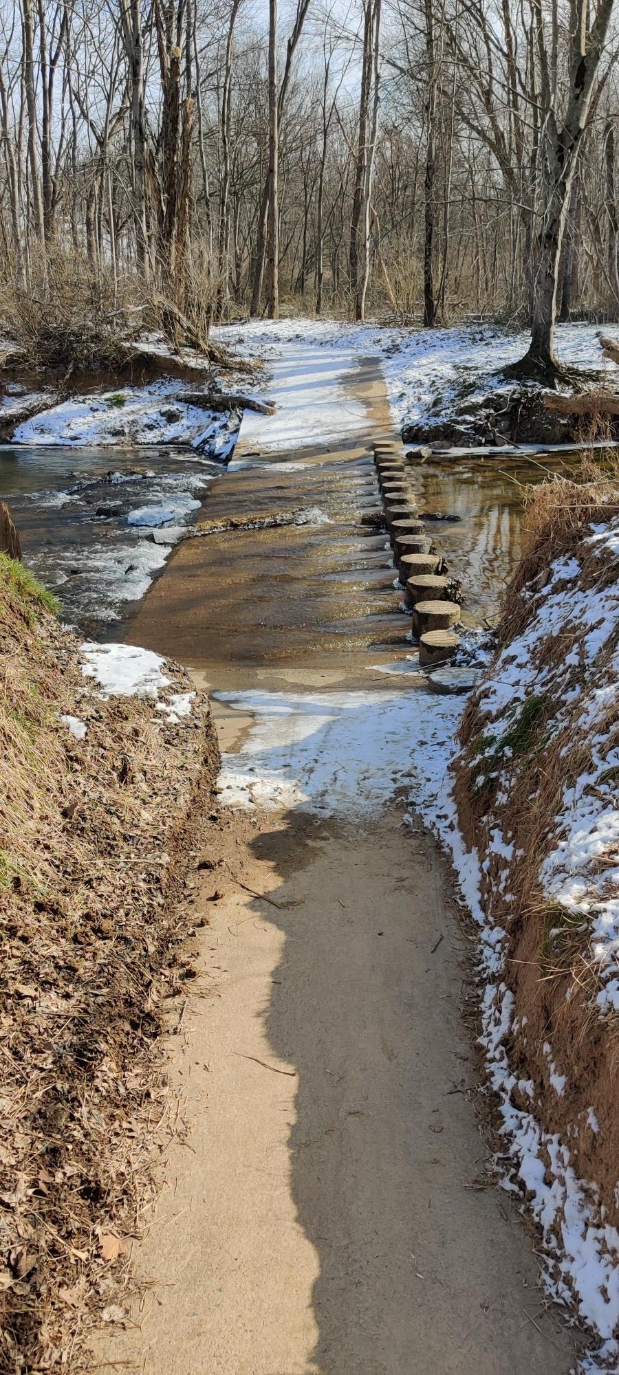 Snowy path leading to a stepping stone bridge over a winter stream.