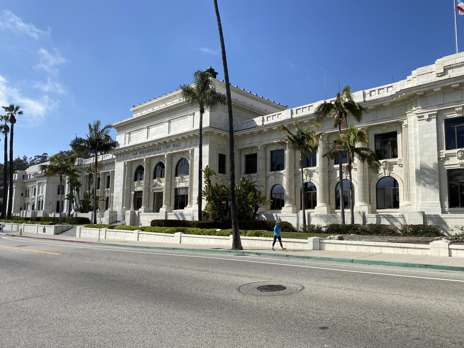 Ornate white building with columns and palm trees; person walking on sidewalk.