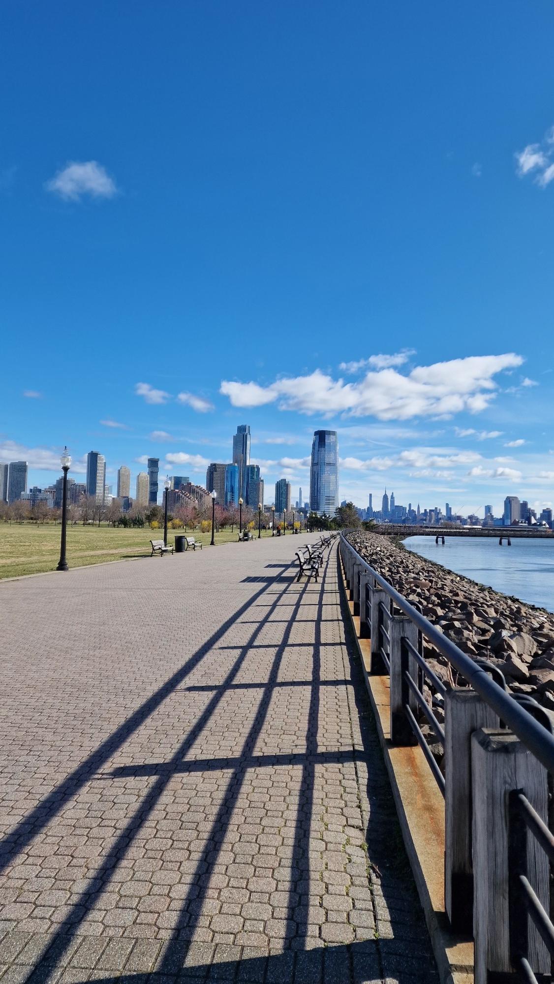 Sunny waterfront promenade with long shadows, city skyline, and blue sky.