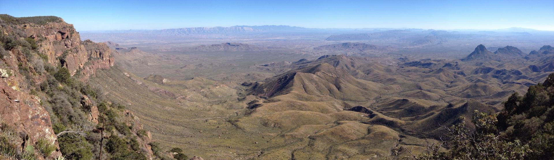 Expansive desert panorama with rugged cliffs, rolling hills, and distant mountains under clear sky.