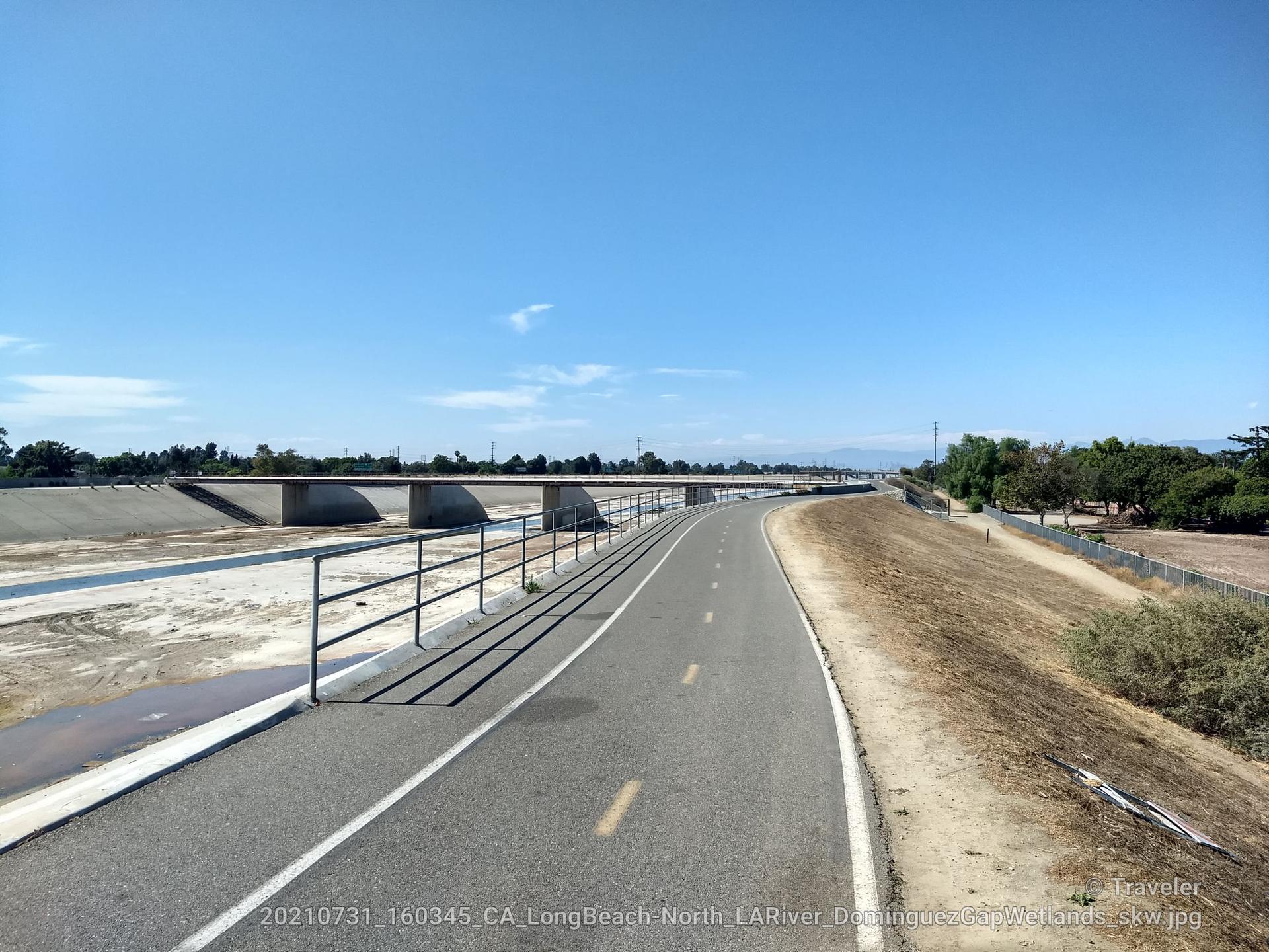 Paved path alongside a wide, dry concrete riverbed with a distant bridge.
