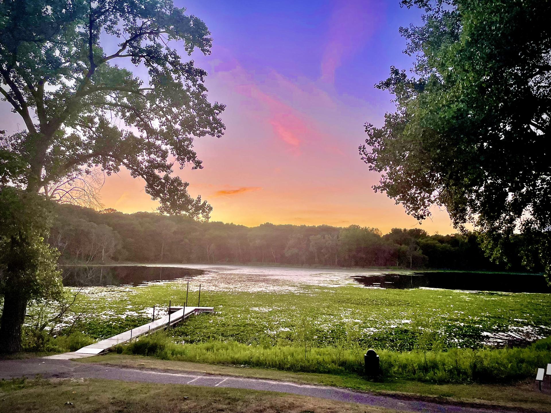A vibrant sunset over a lake with lily pads and a wooden dock.