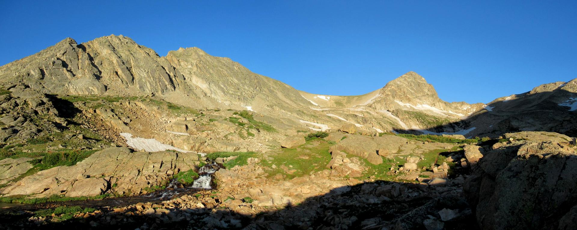 Sunny mountain valley with rocky peaks, a stream, and green patches.