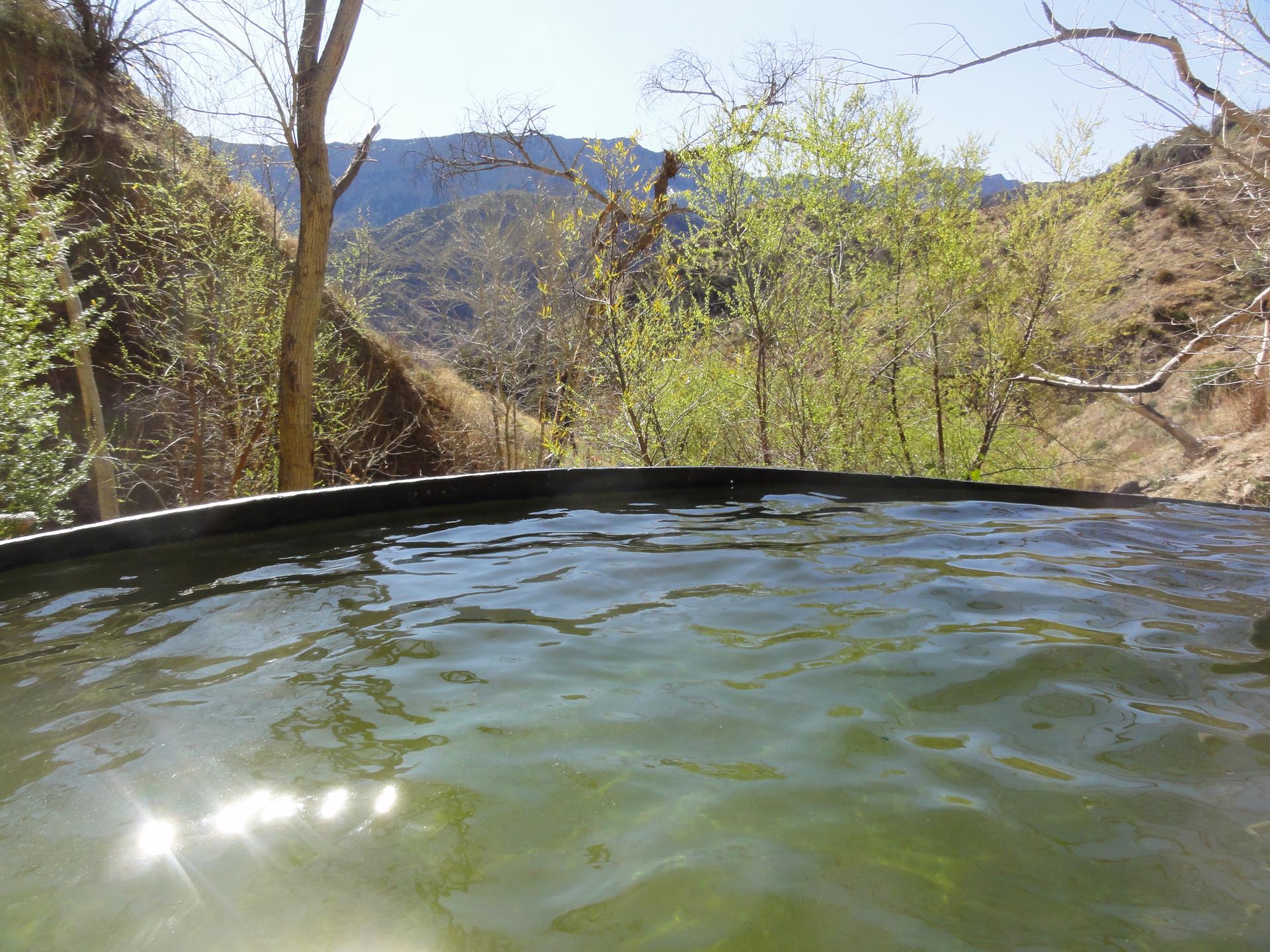 Sunlight glints on a natural pool with green trees and mountains beyond.