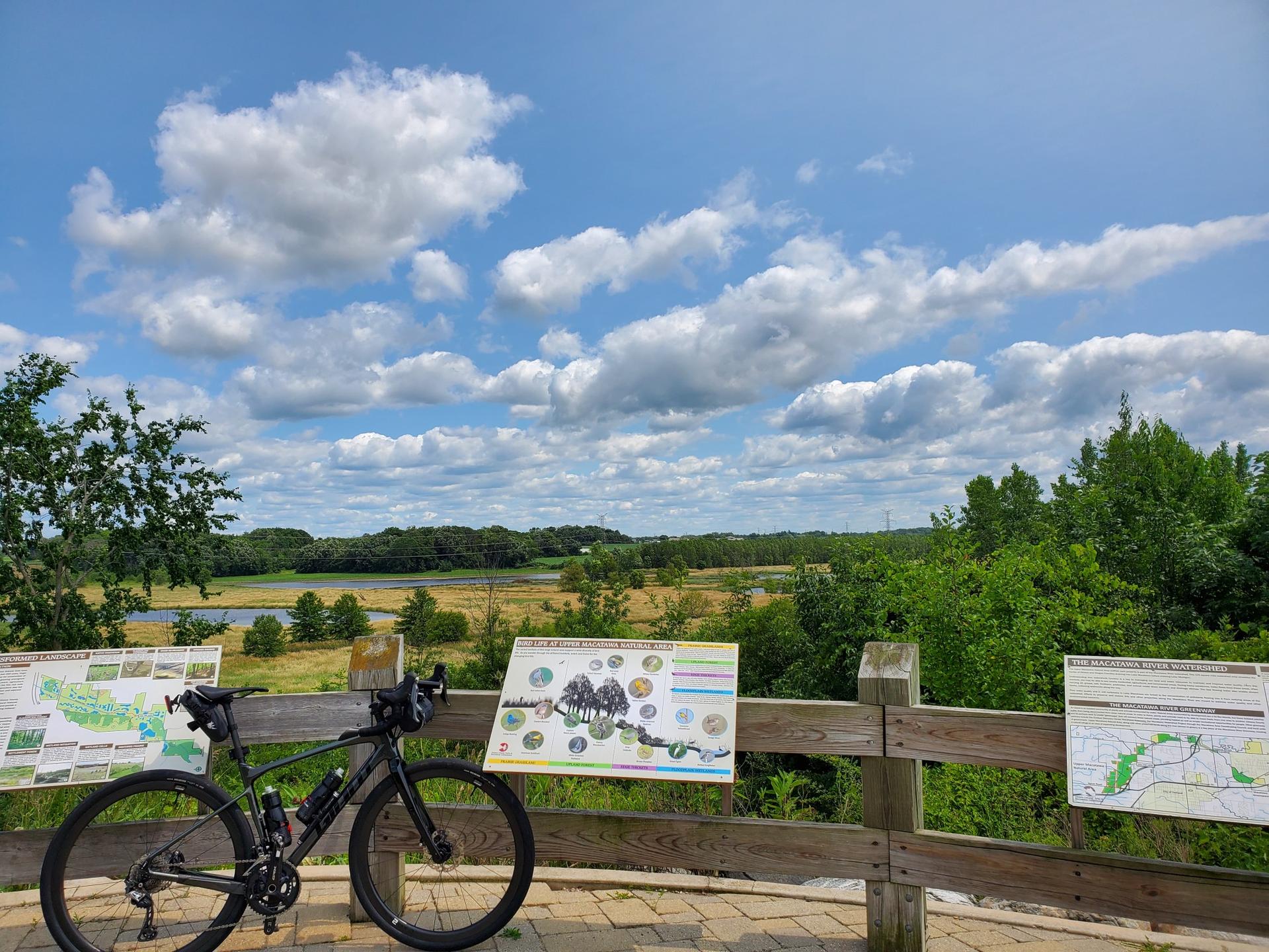 Bicycle parked at an overlook with signs, overlooking a sunny landscape with water.