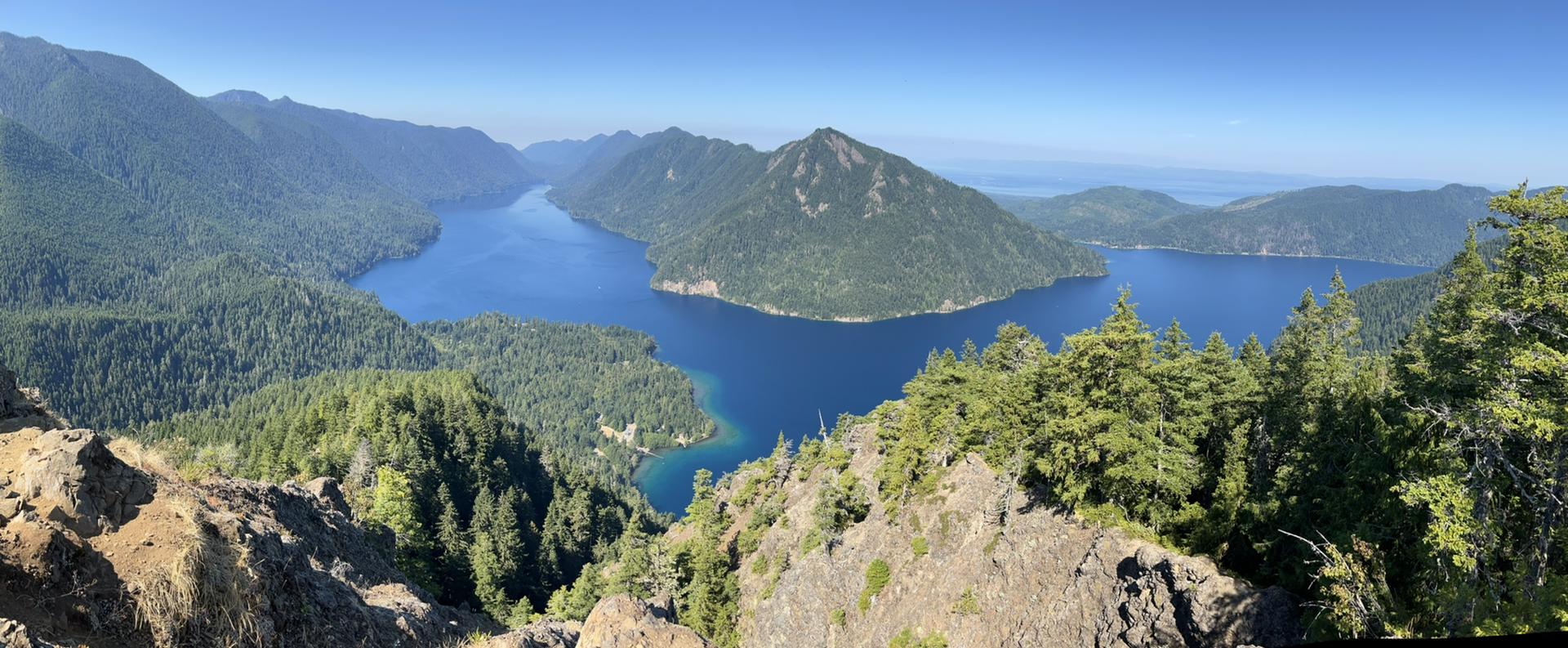 Panoramic view of a deep blue lake surrounded by lush green mountains under clear skies.