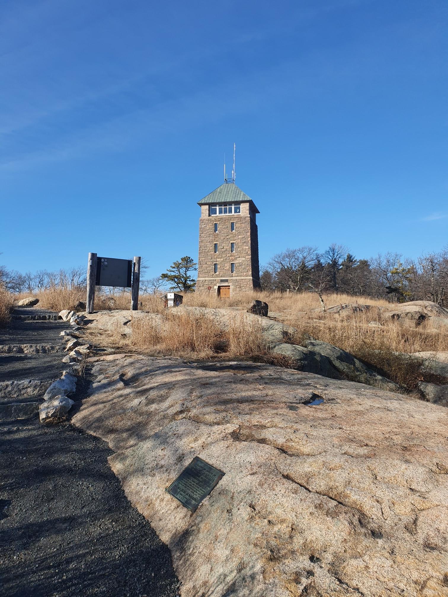 A stone tower stands on a rocky, grassy hilltop under a bright blue sky.