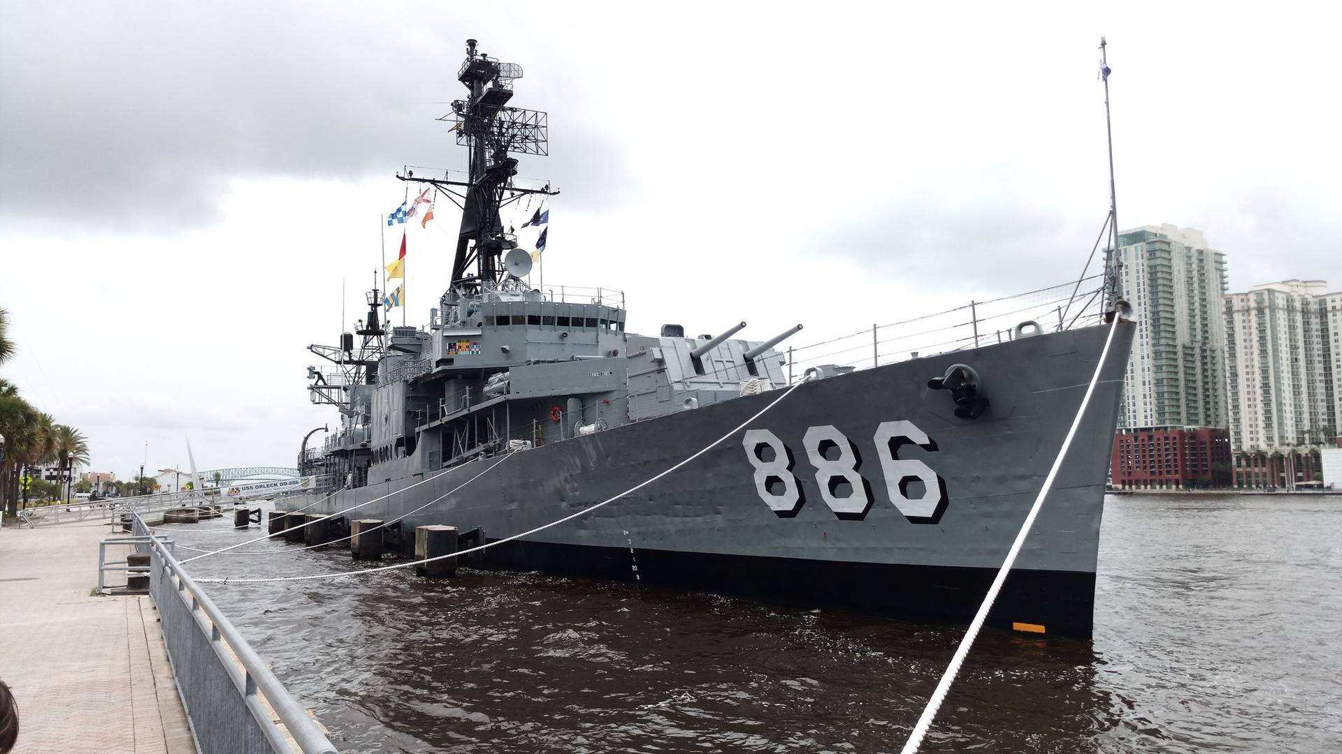 Large gray warship 886 docked at a pier with city buildings behind.