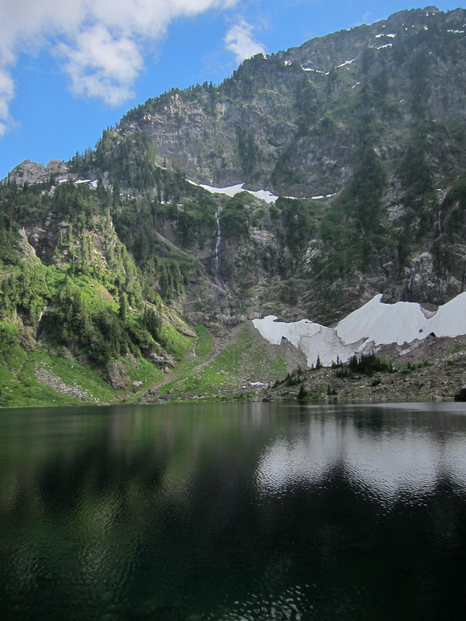 Serene lake reflecting green and rocky mountains under a bright blue sky.
