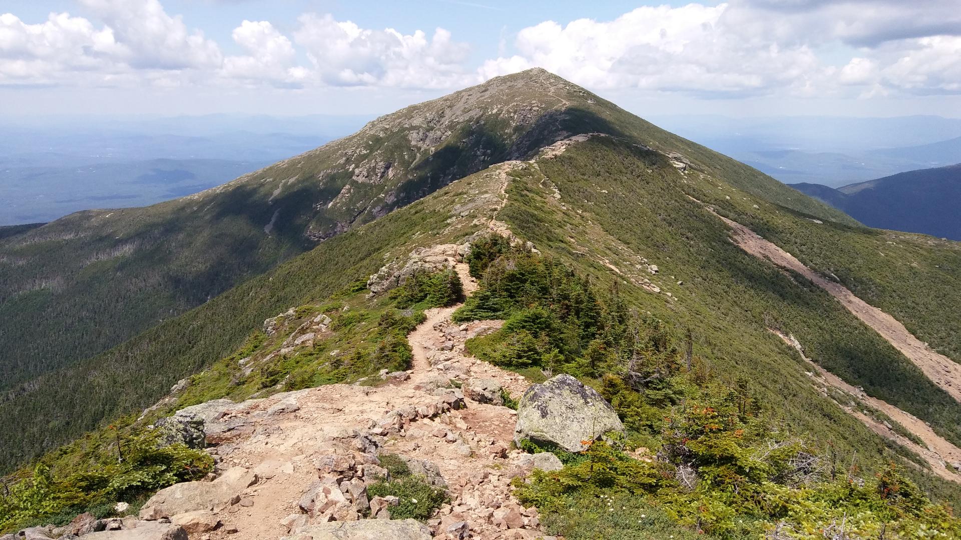 A rugged, rocky mountain ridge leads to a distant peak under a bright sky.