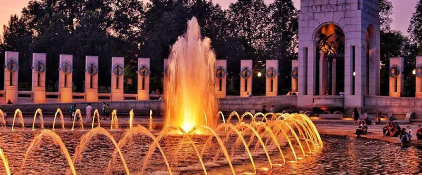 Illuminated fountains at dusk, archway and columns, people sitting.