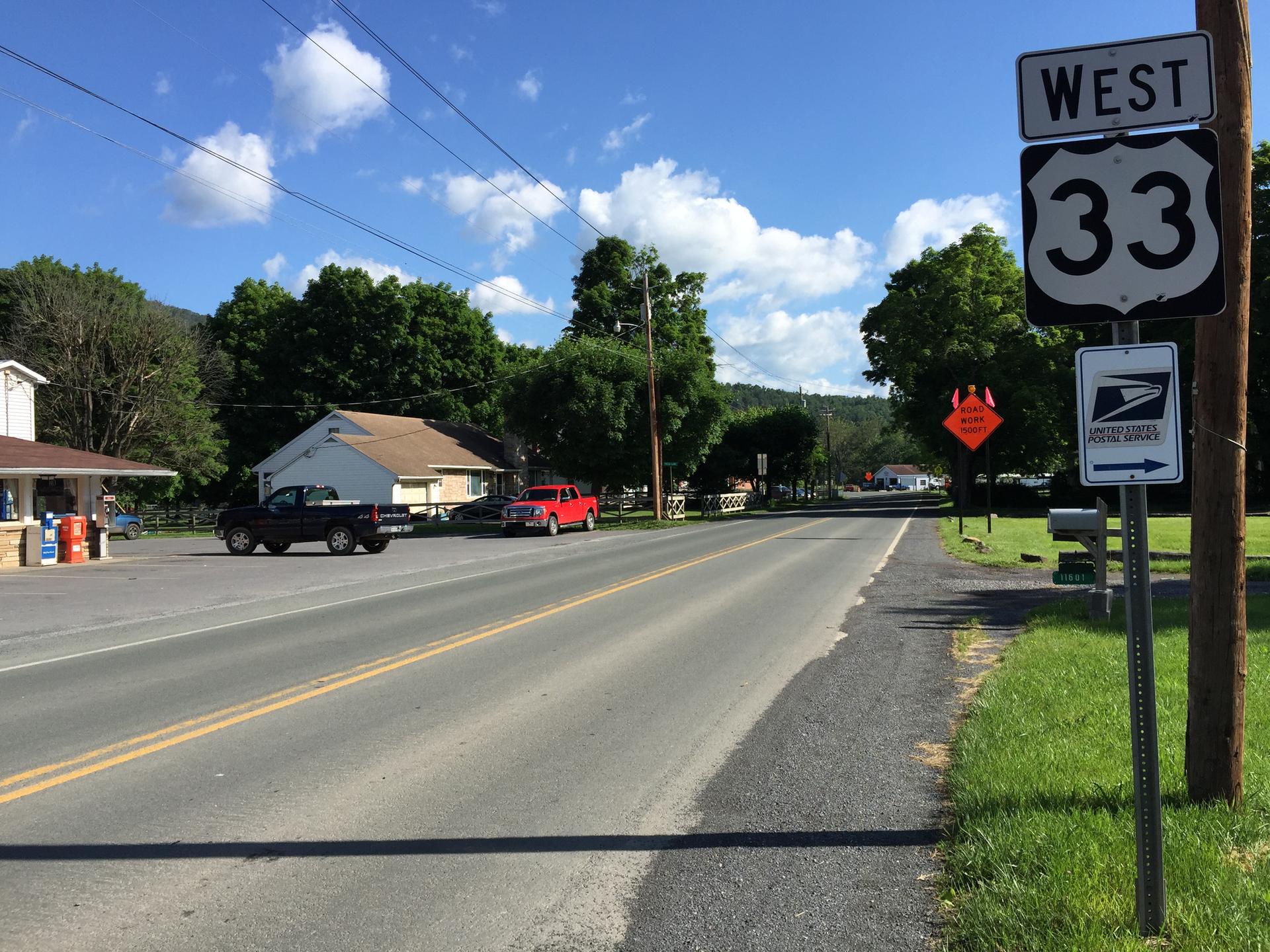 Rural road with signs, houses, and distant mountains under blue sky.