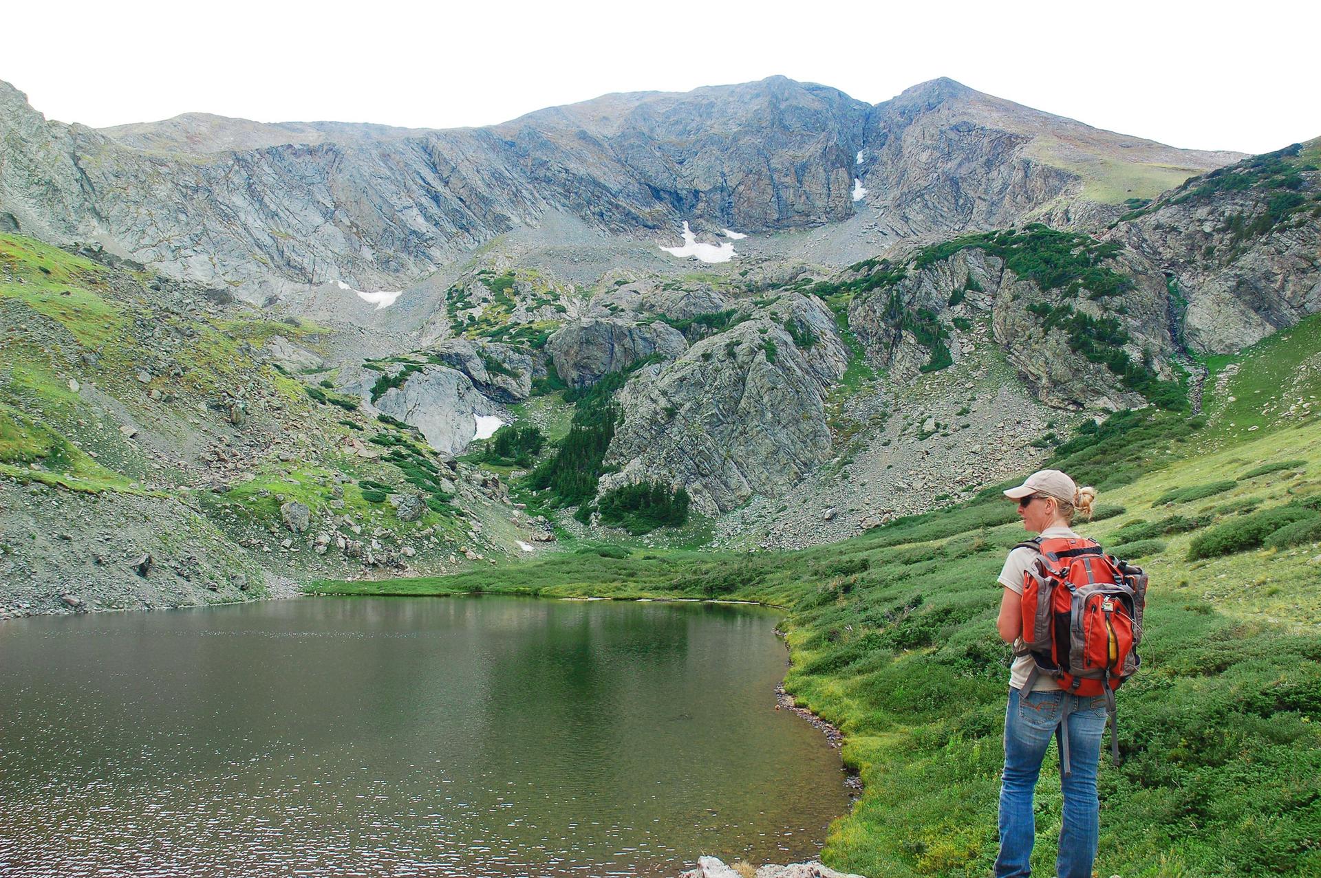 Hiker with backpack stands by a mountain lake under an overcast sky.