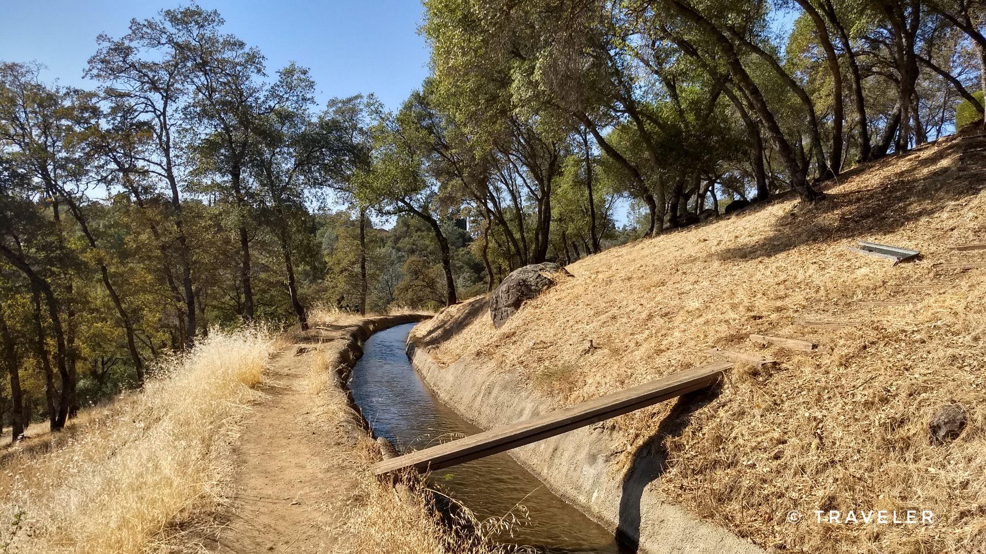 Sunny view of a concrete water channel, dirt path, and dry grassy hill.