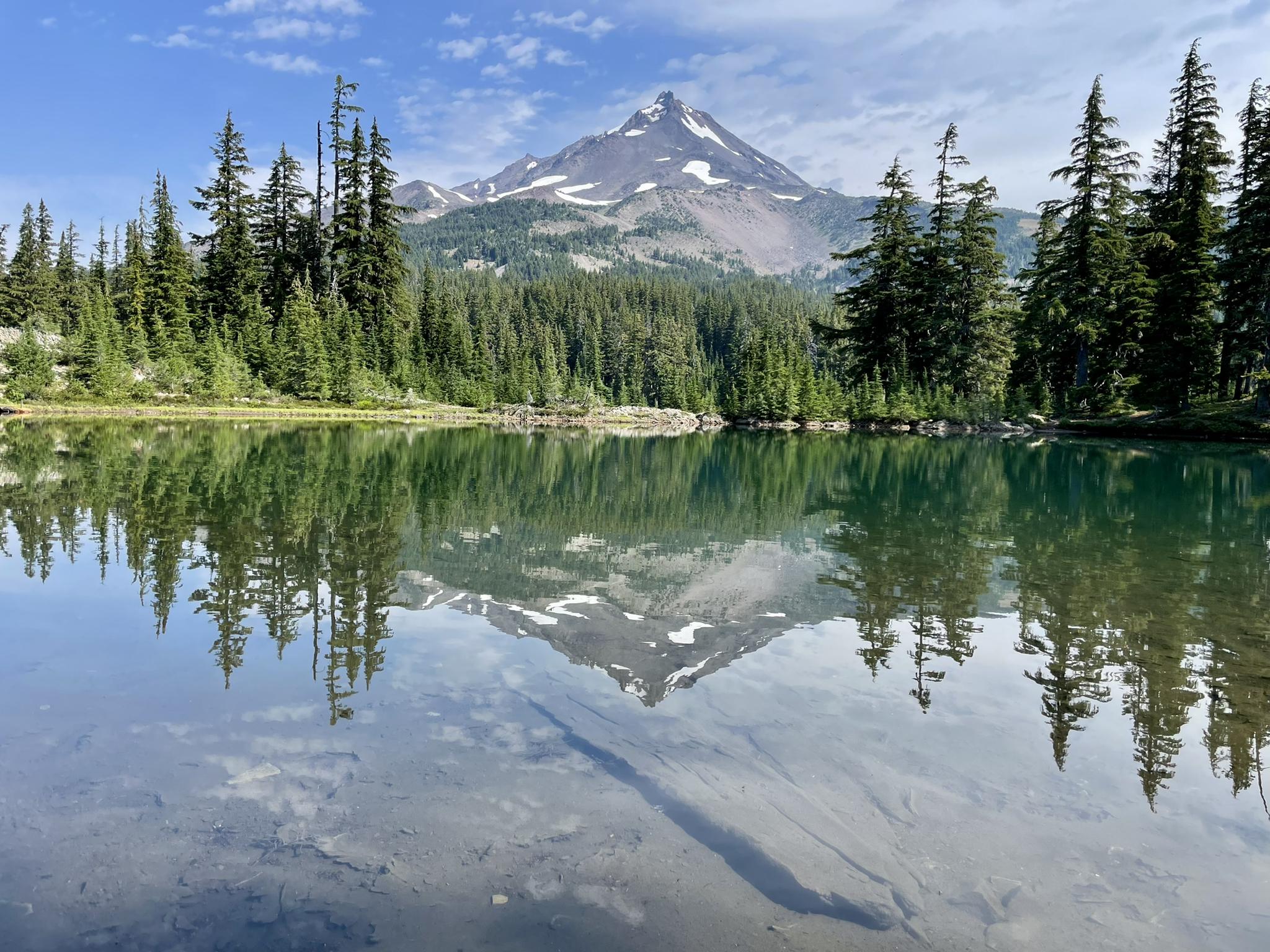 Snow-capped mountain reflecting perfectly in a calm, clear lake with pine trees.