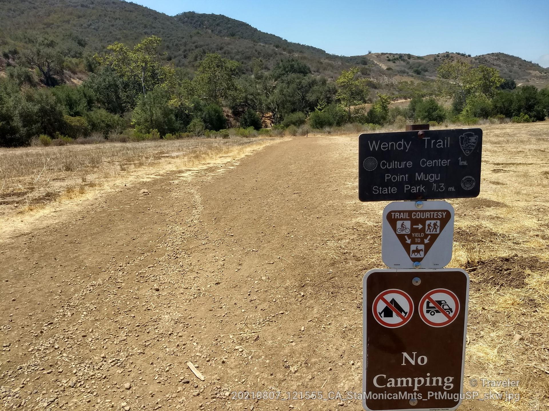 Dirt trail with trail signs, dry grass, and distant hills under clear sky.