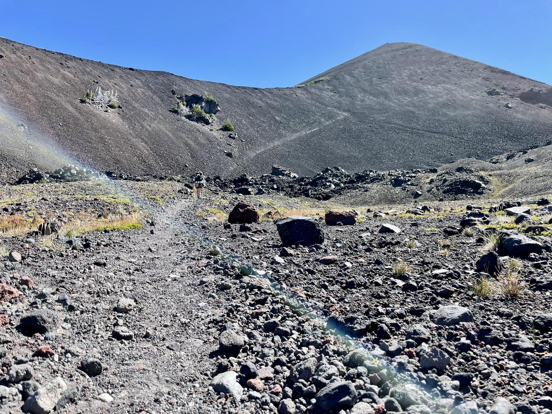 A rocky path ascends a dark, barren volcanic landscape under a bright sky.
