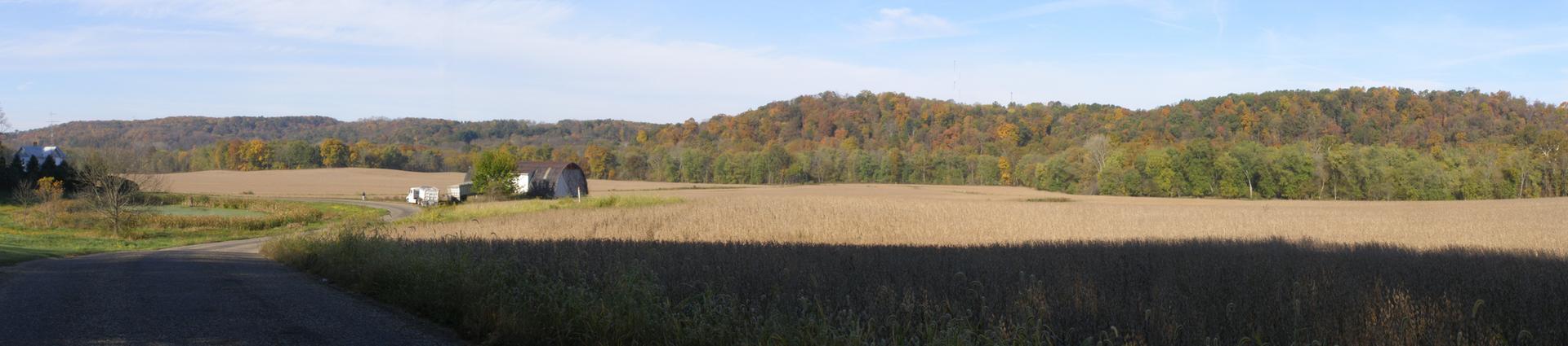 Panoramic view of harvested fields, small buildings, and distant wooded hills.