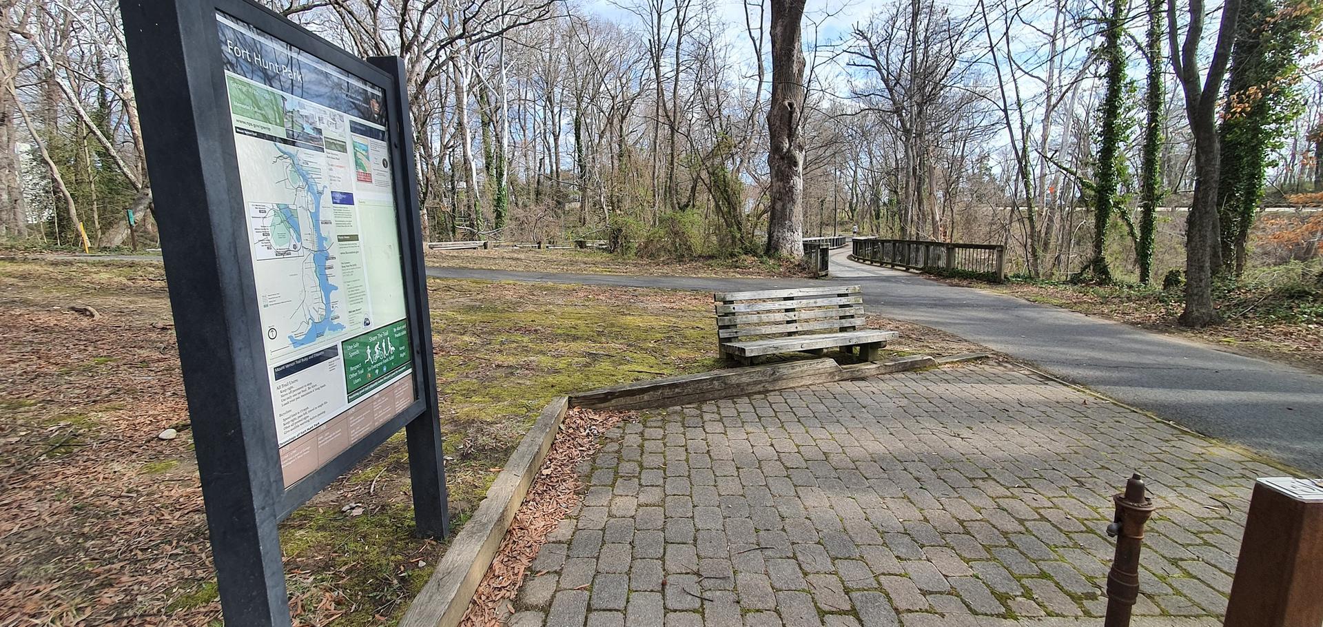 Park sign and wooden bench next to a paved path in a wooded area.