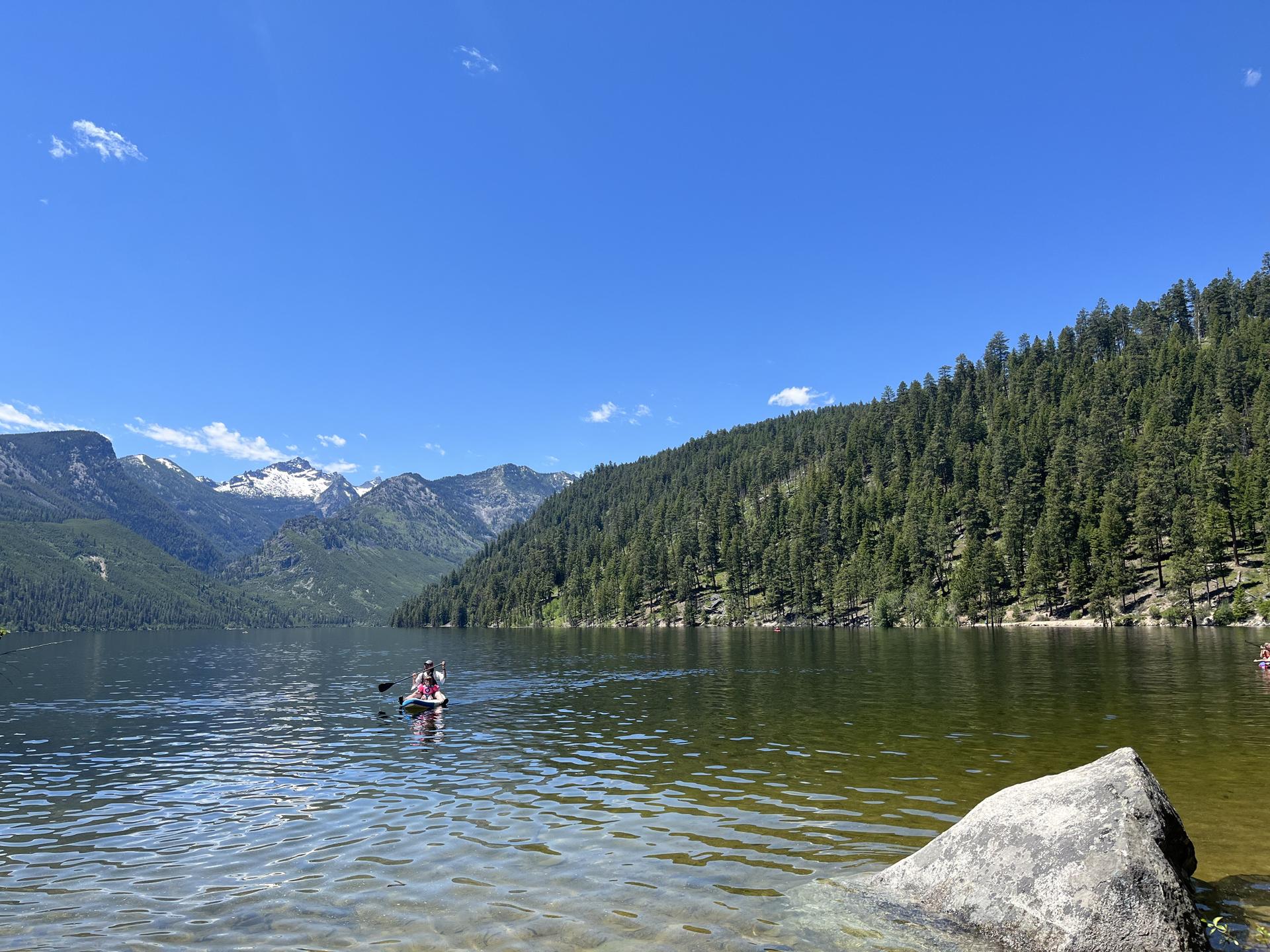 Person paddleboarding on a clear lake surrounded by forested mountains under a blue sky.