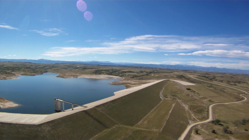 Large reservoir and dam with dry hills under a bright, partly cloudy sky.