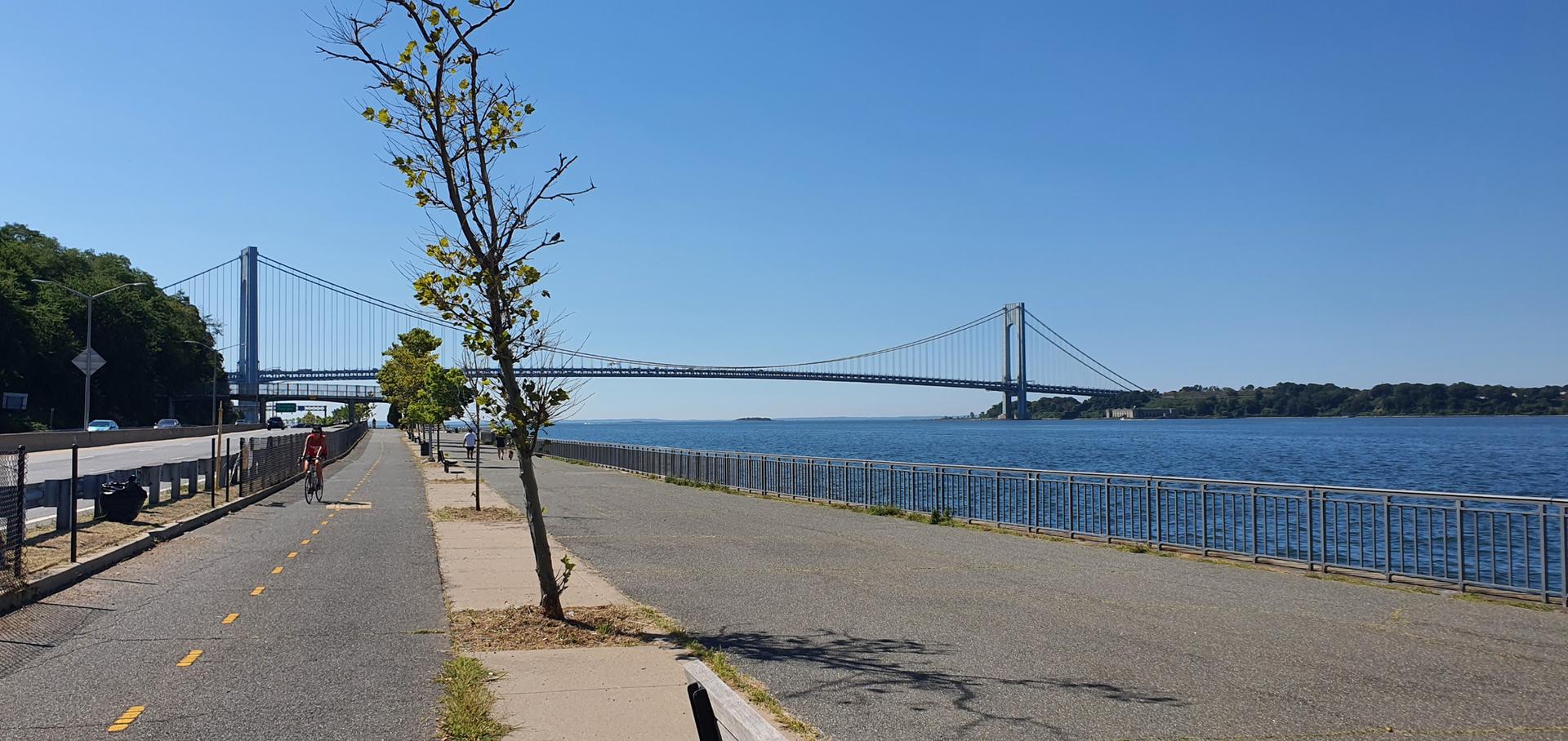 Long suspension bridge over wide water next to a sunny paved path.