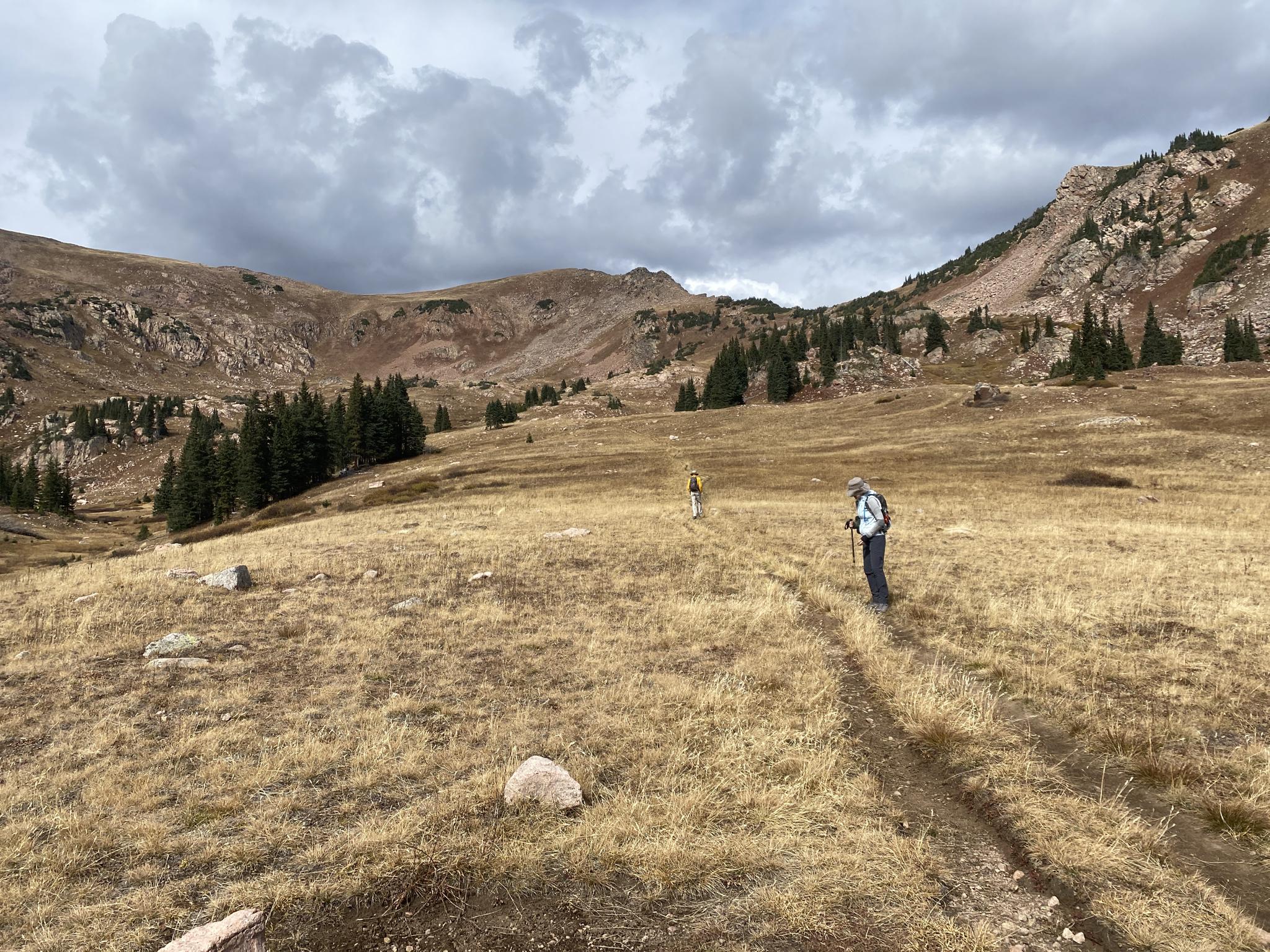Two hikers on a dirt trail in a vast, cloudy mountain valley.