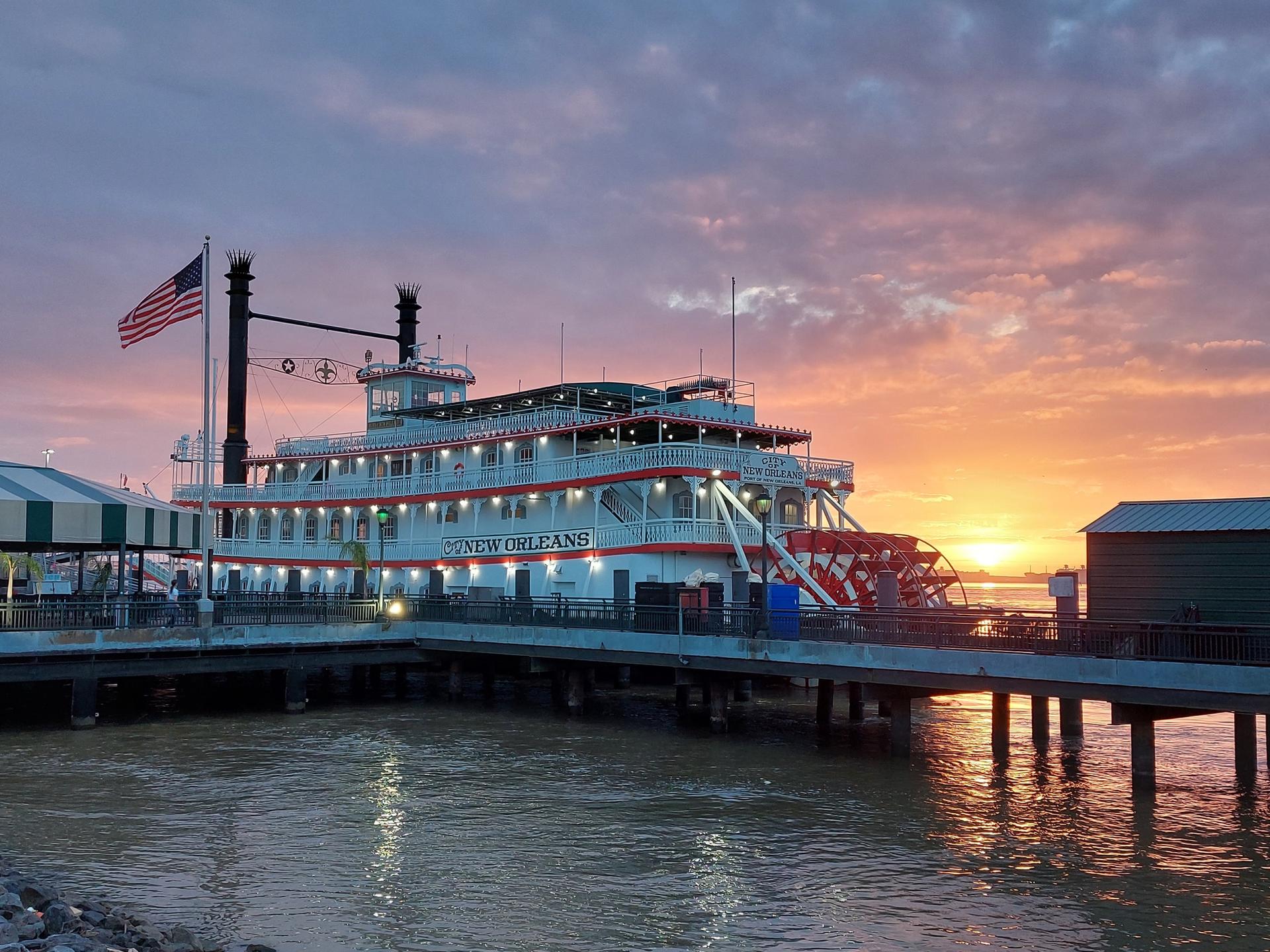Paddlewheel boat docked at sunset, with vibrant orange and pink sky.