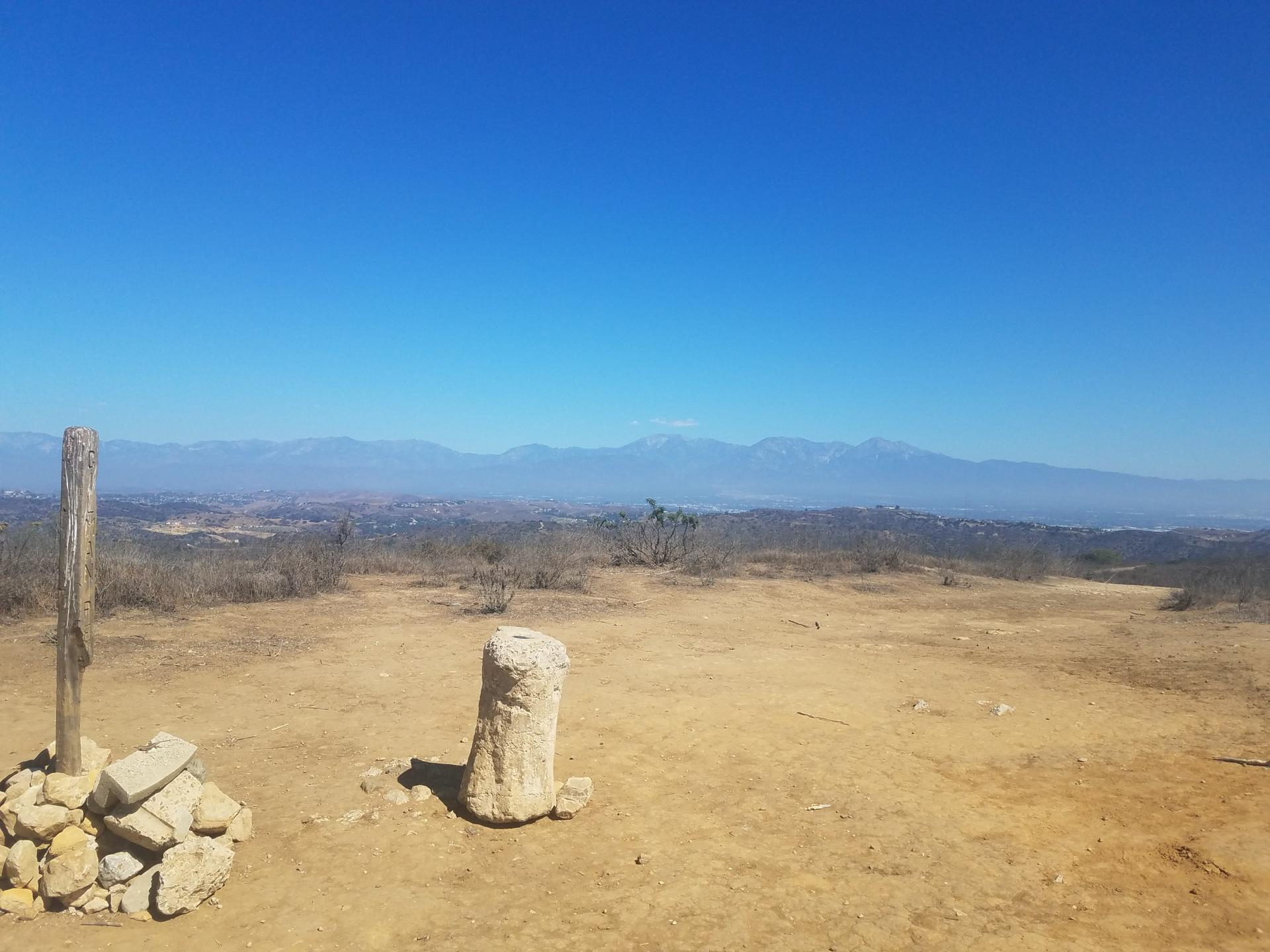 Arid hilltop with wooden post, stone marker, and distant mountains under clear sky.